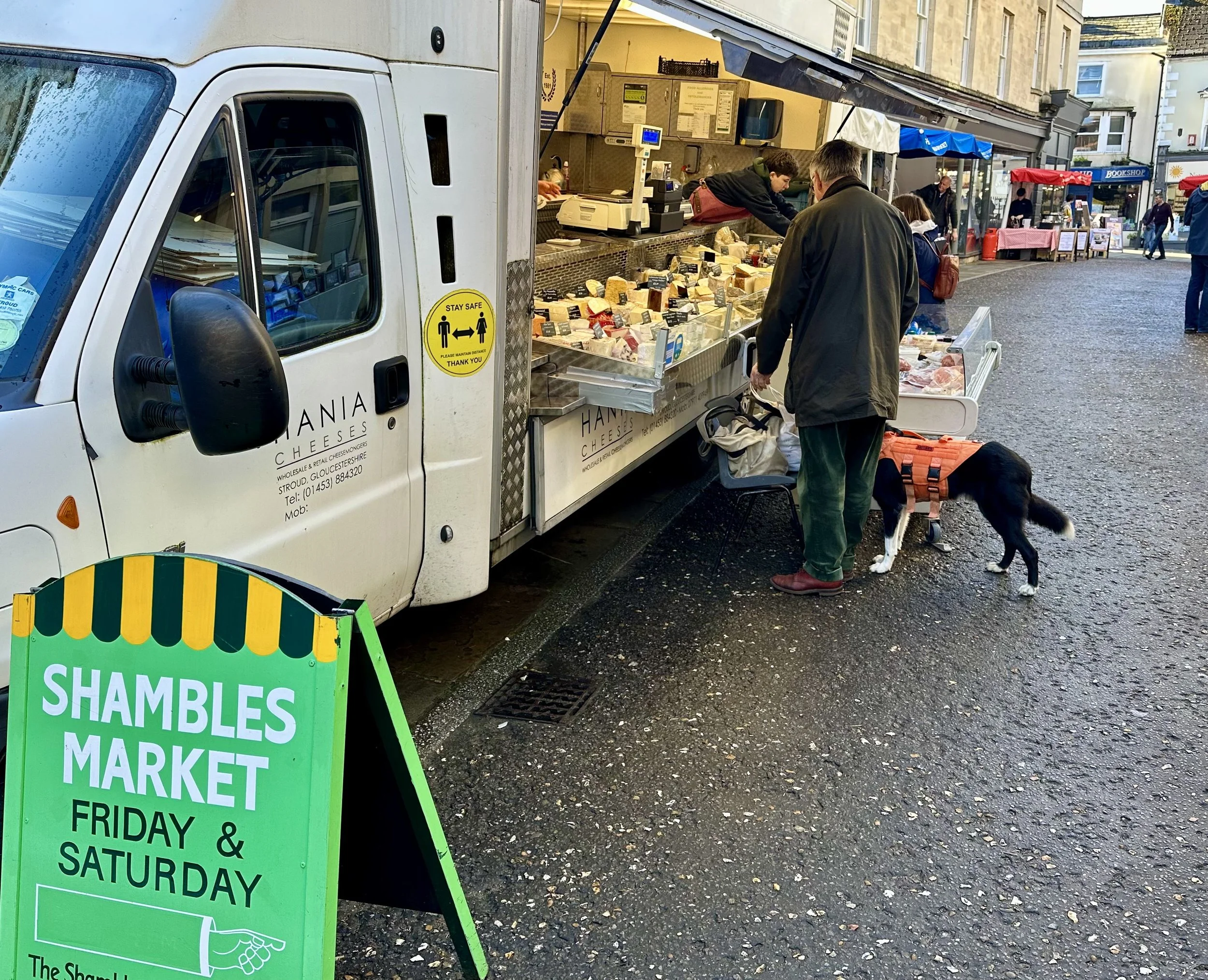 A street market scene with a cheese truck selling cheese products. People are browsing the cheese selection. A green sign reads "Shambles Market, Friday & Saturday." It is a cloudy day.