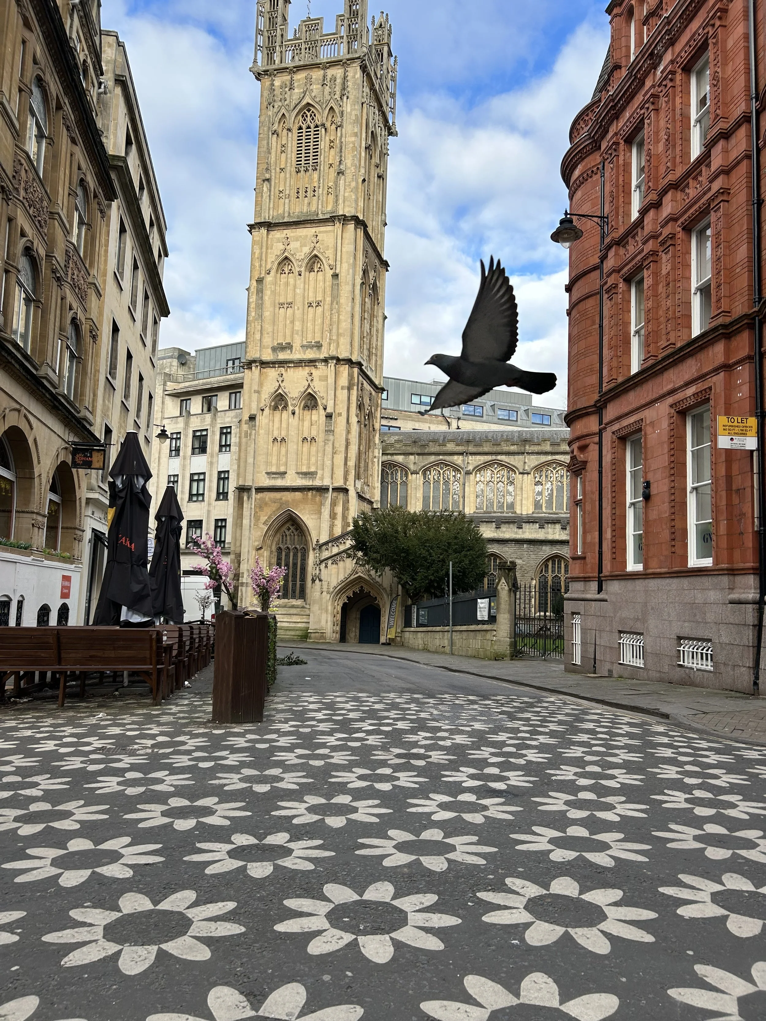 Empty city street with patterned flower design on the pavement, historic buildings on two sides, a prominent church tower in the background, pink flowering trees, outdoor seating area, and a bird flying overhead.