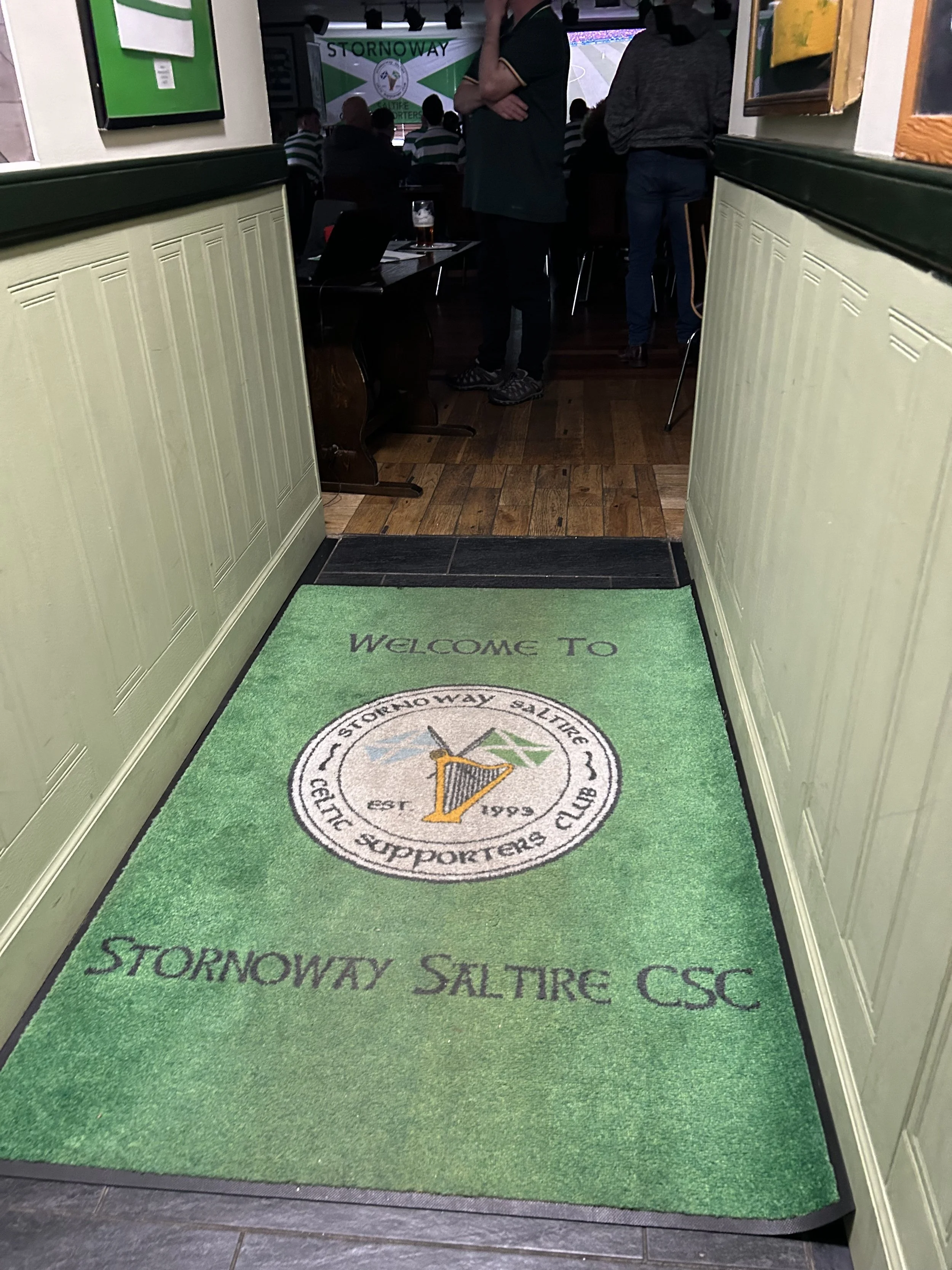 Green welcome mat with the logo and text of the Stornoway Saltire Supporters Club, placed at the entrance to a room where people are gathered, some standing and watching a sports game on a TV screen.