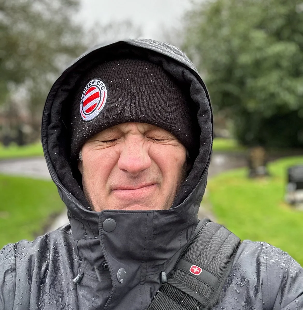 A man outdoors wearing a black beanie with a logo, a black waterproof jacket, and a backpack, squinting with a serious expression on a rainy day.