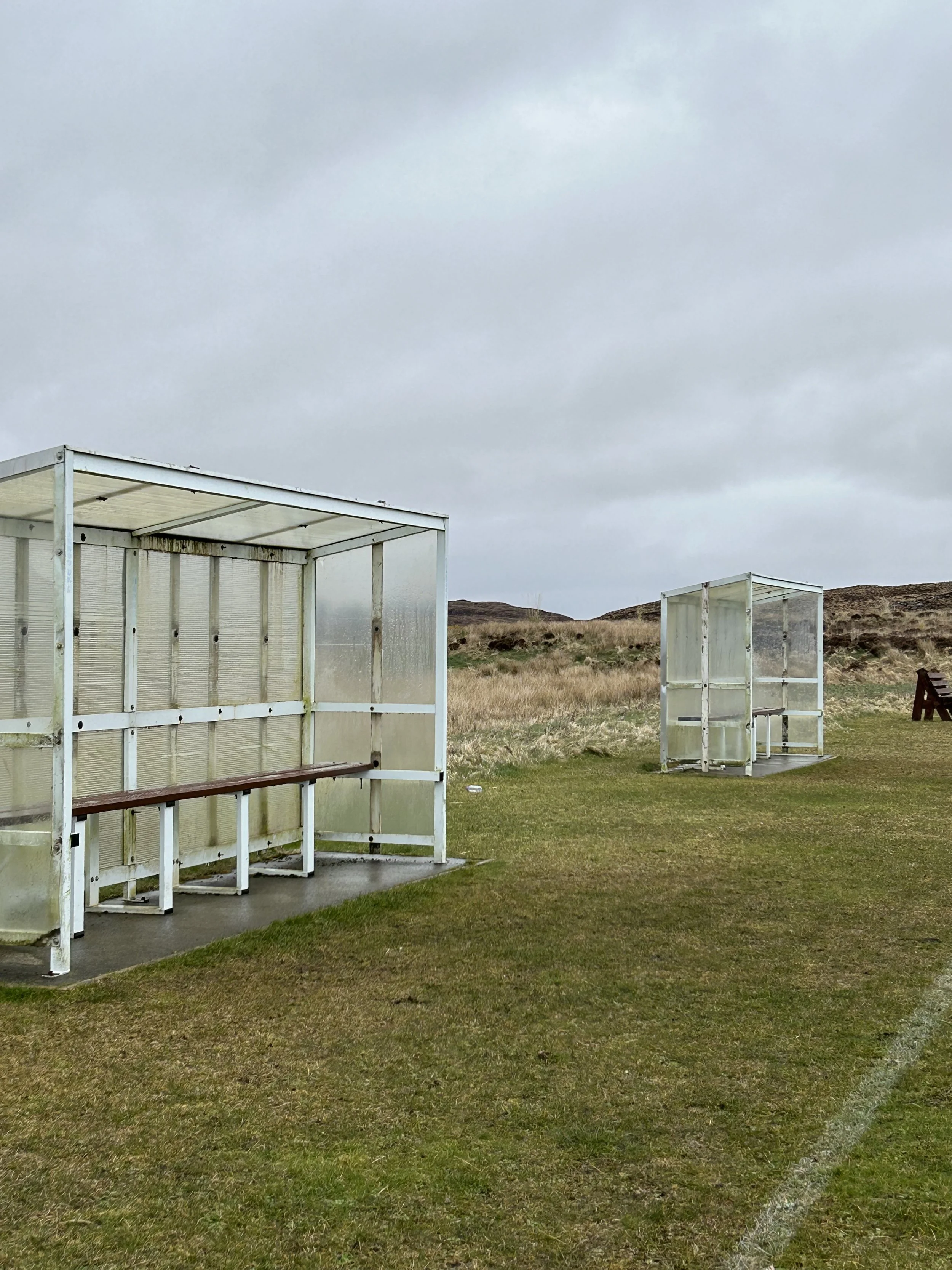 Two small, transparent bus stops on grassy field with cloudy sky in background.