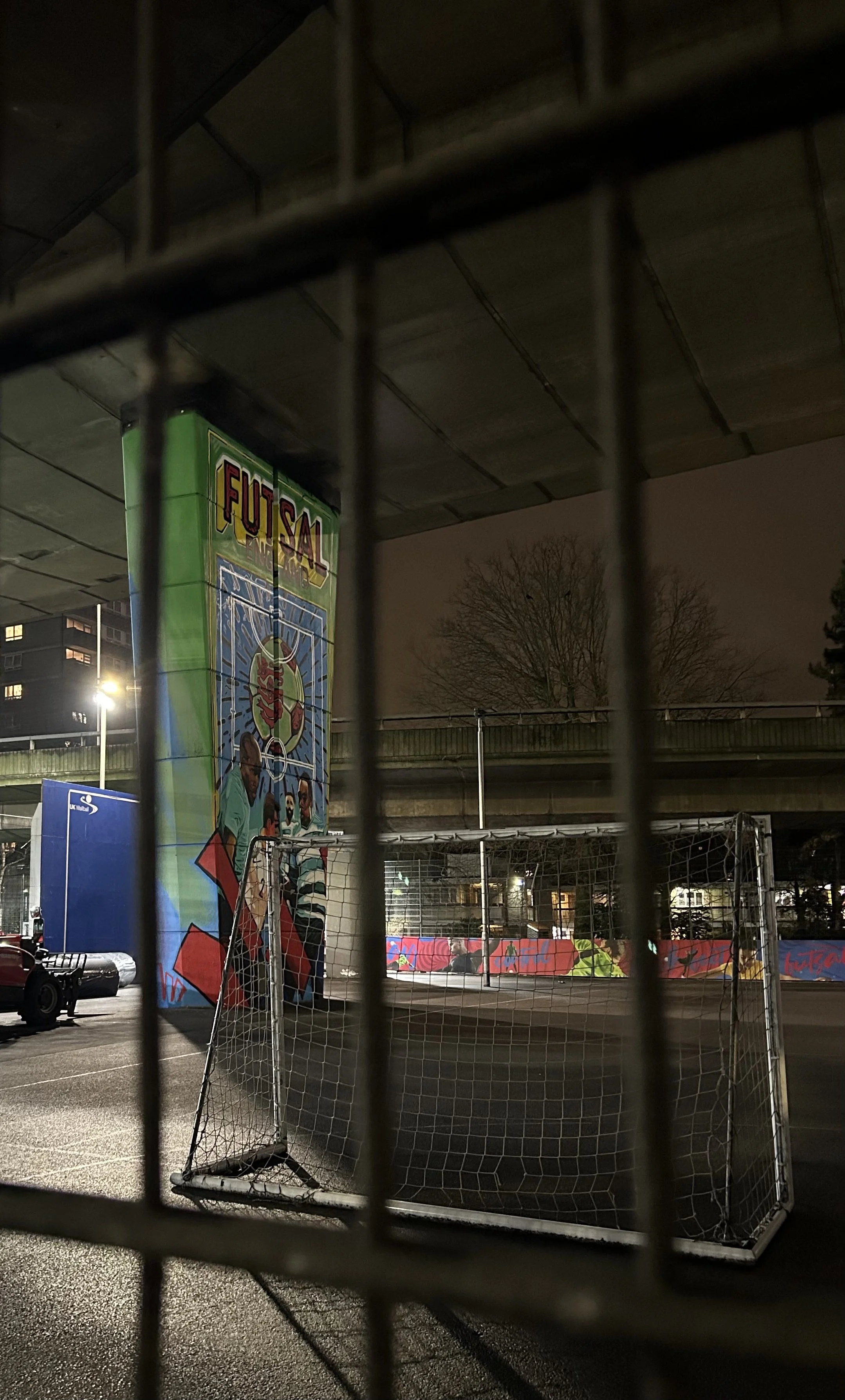 Nighttime view of an urban sports court surrounded by a fence, featuring a mural with the word 'FUTSAL' and images of people, under an overpass with leafless trees in the background.