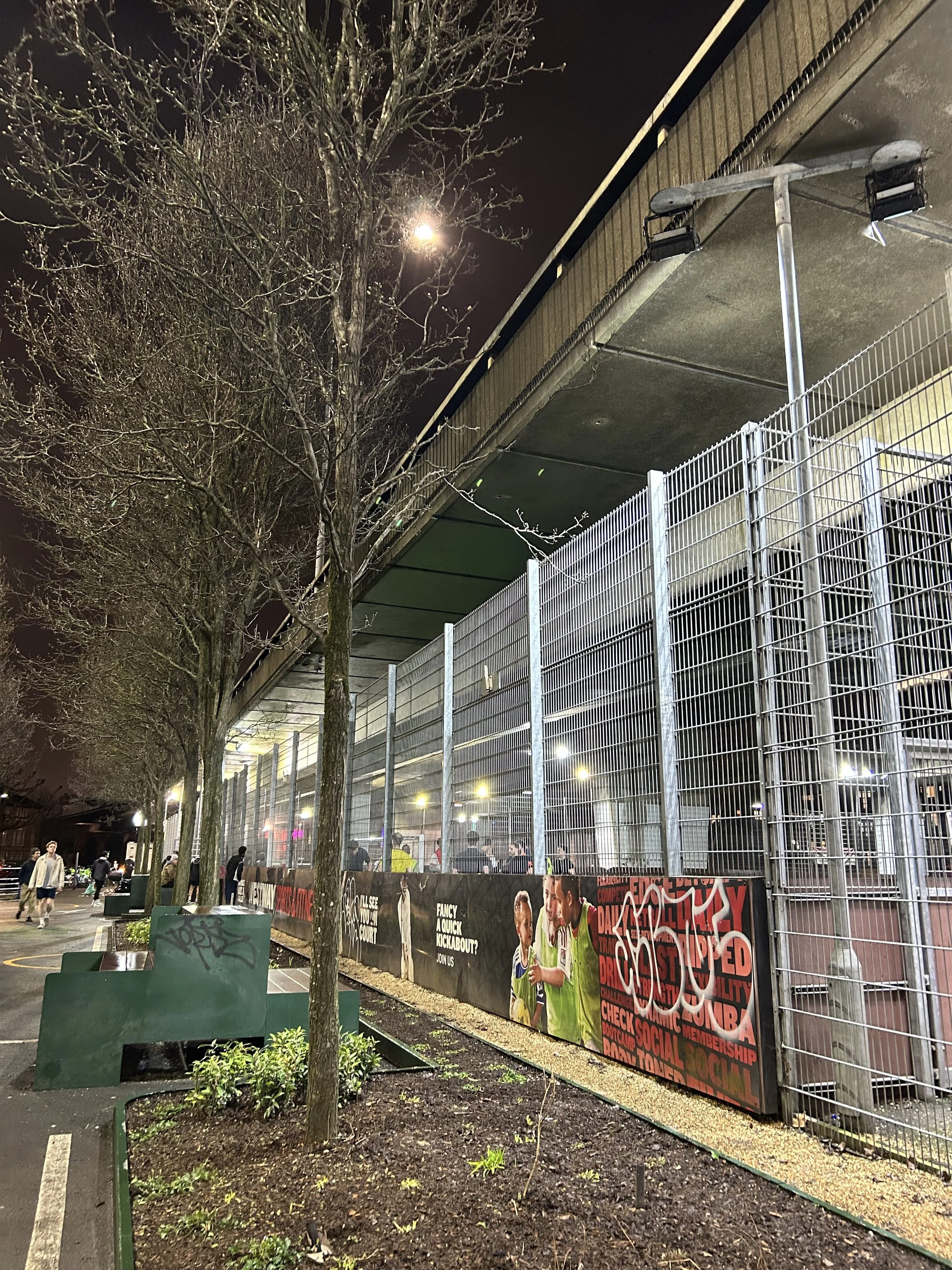 Nighttime street scene showing a parking area with a row of leafless trees, green benches, and a fence with social media graffiti and advertisements along a building with a large overpass above.