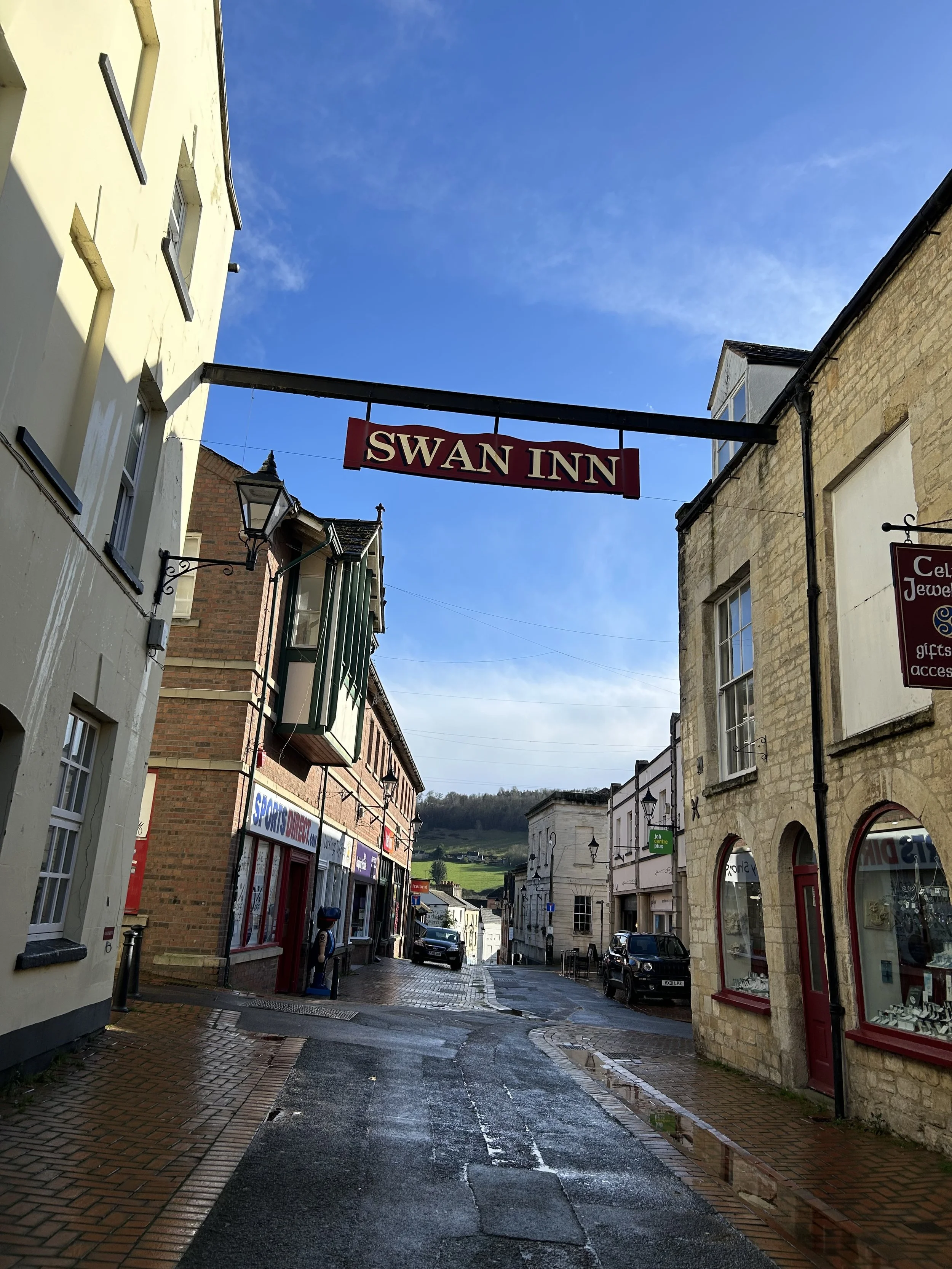 A street view in a small town with a sign for Swan Inn hanging above the road, showing buildings, cars, and a hill in the background under a partly cloudy sky.