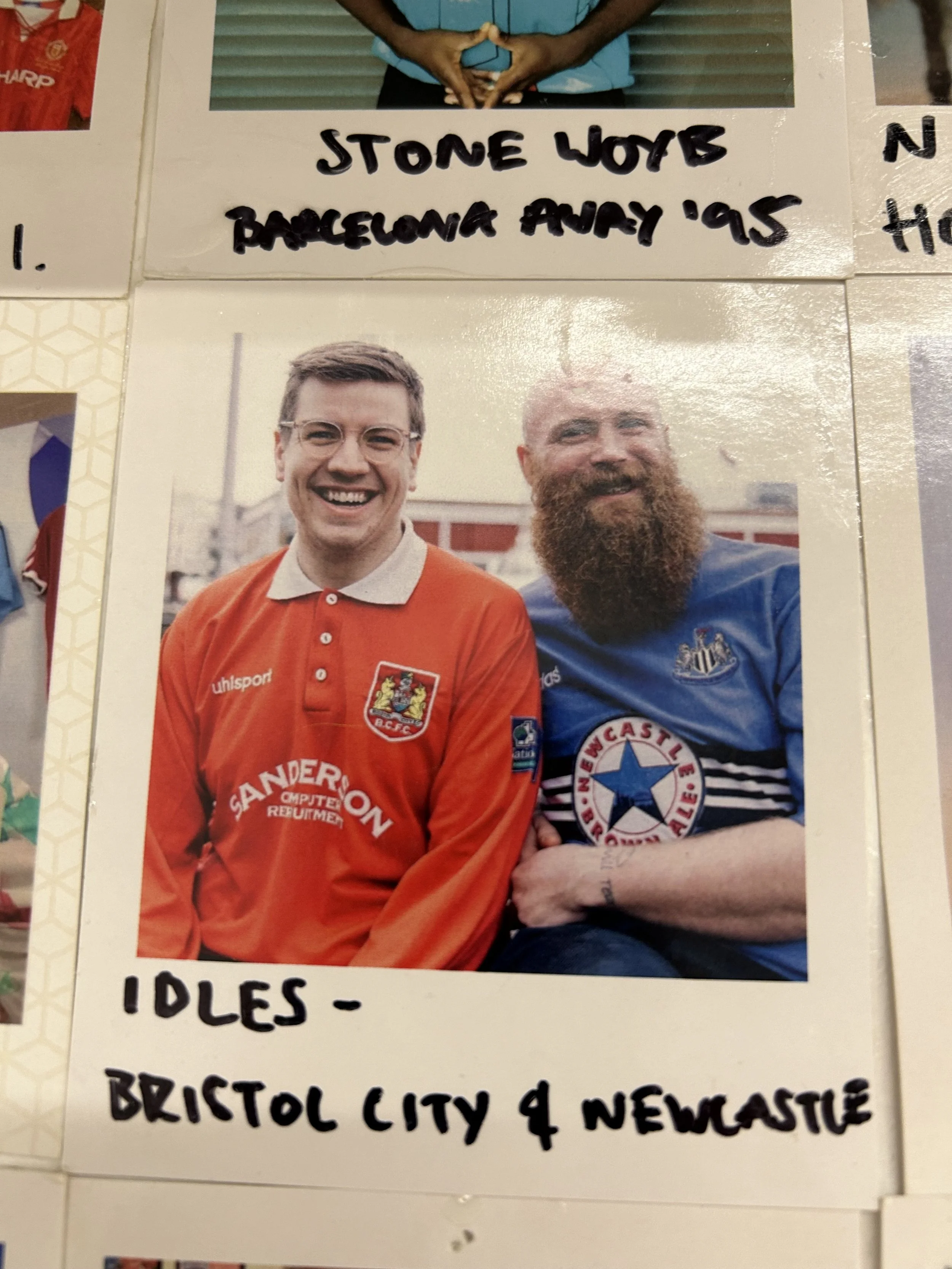 Two men smiling, one wearing a red Bristol City soccer jersey and the other wearing a blue Newcastle United soccer jersey, sitting outdoors in daylight.