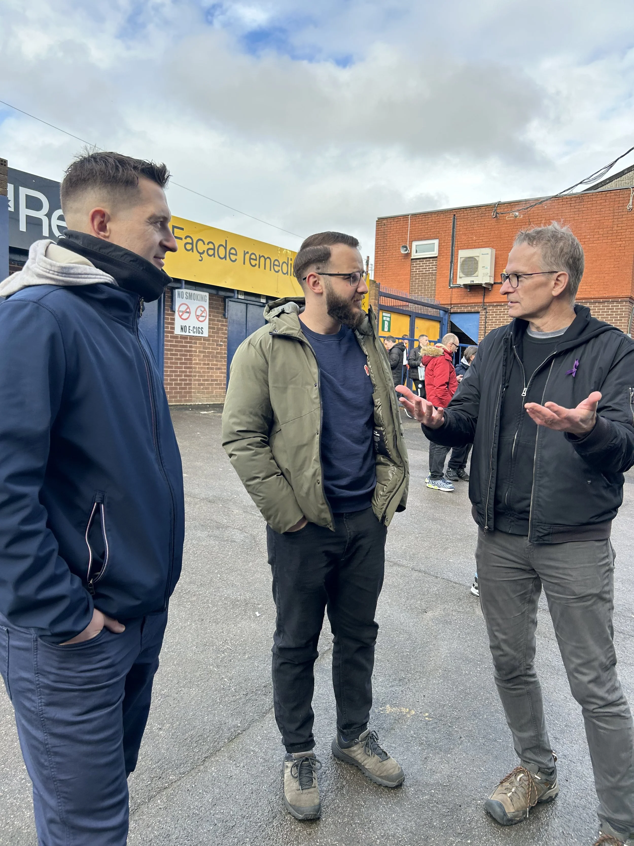 Three men having a conversation outdoors near a building with a yellow sign that reads 'Façade remedial'.