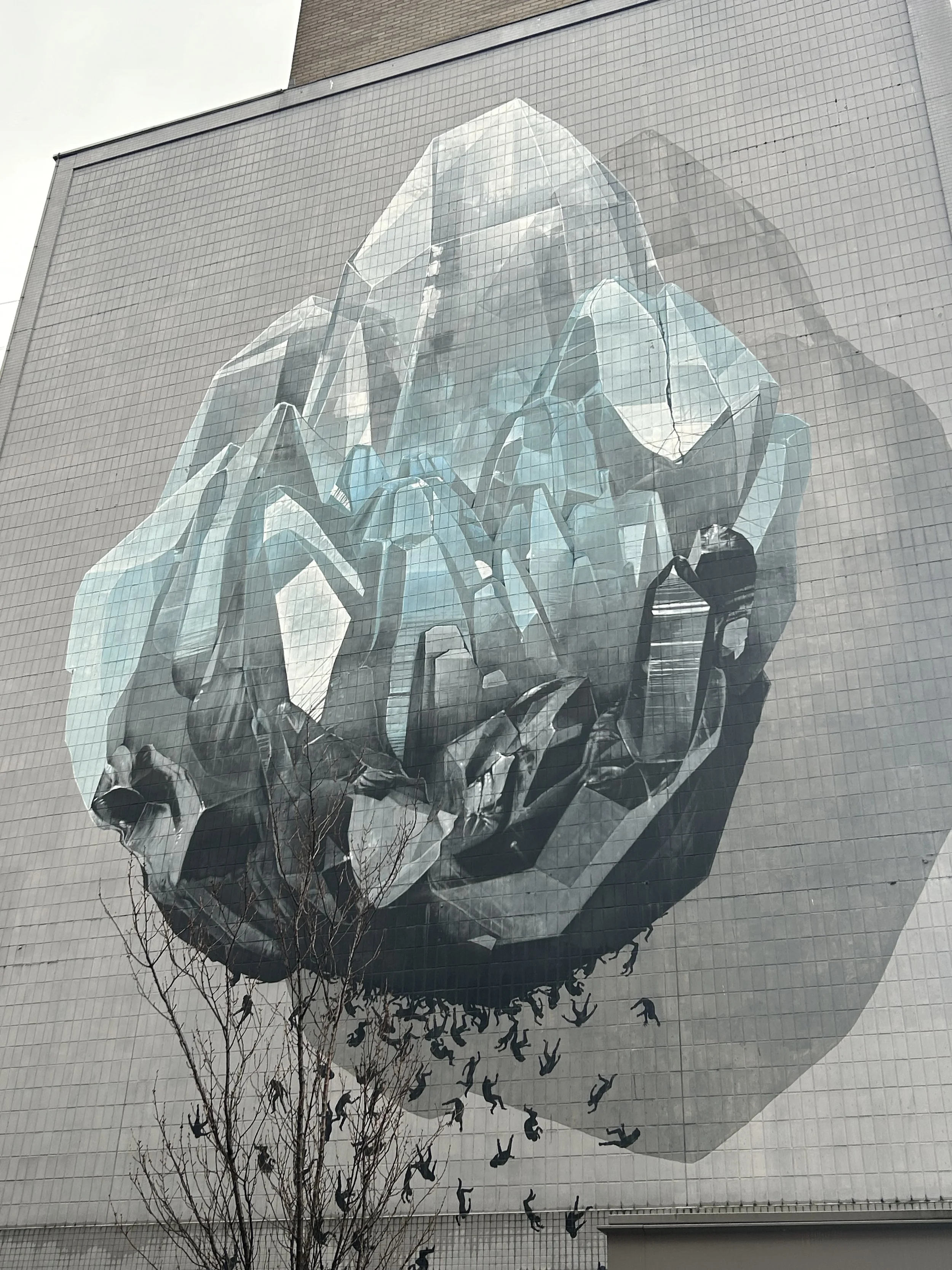 A large mural on a building depicting a stylized, abstract human brain with reflective, glass-like surfaces. Small black silhouettes of people are shown falling or flying downward from the brain.