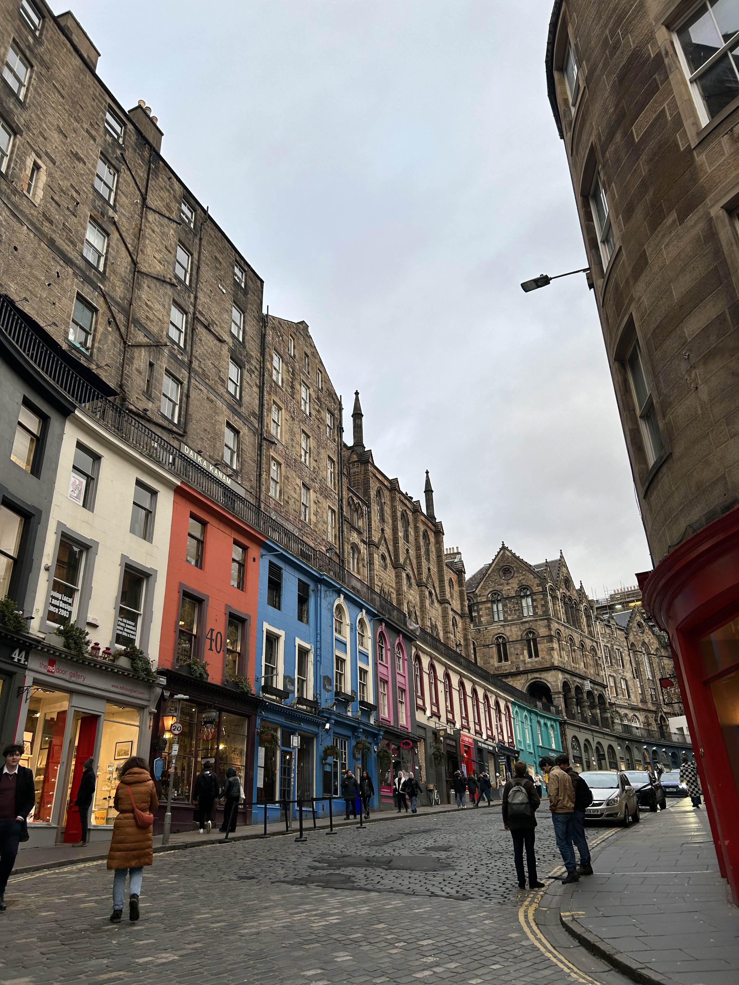 Colorful shops on a cobblestone street with people walking and cars parked on the side, under a gray sky.