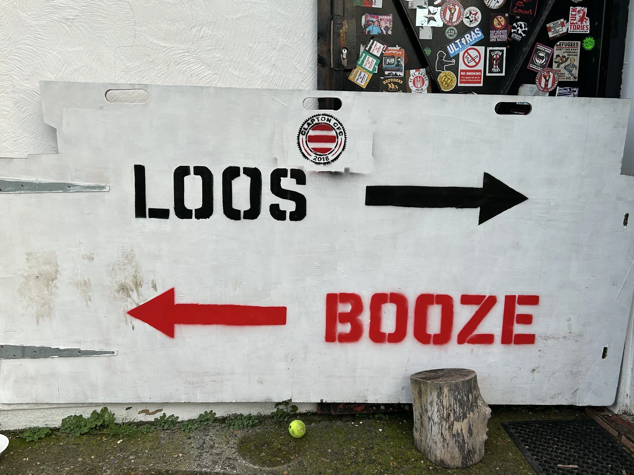 White sign with black and red arrows and text reading "LOOS" pointing right and "BOOZE" pointing left, outdoors on a wall with stickers and graffiti, a tennis ball, a tree stump, and a doormat visible at the bottom.