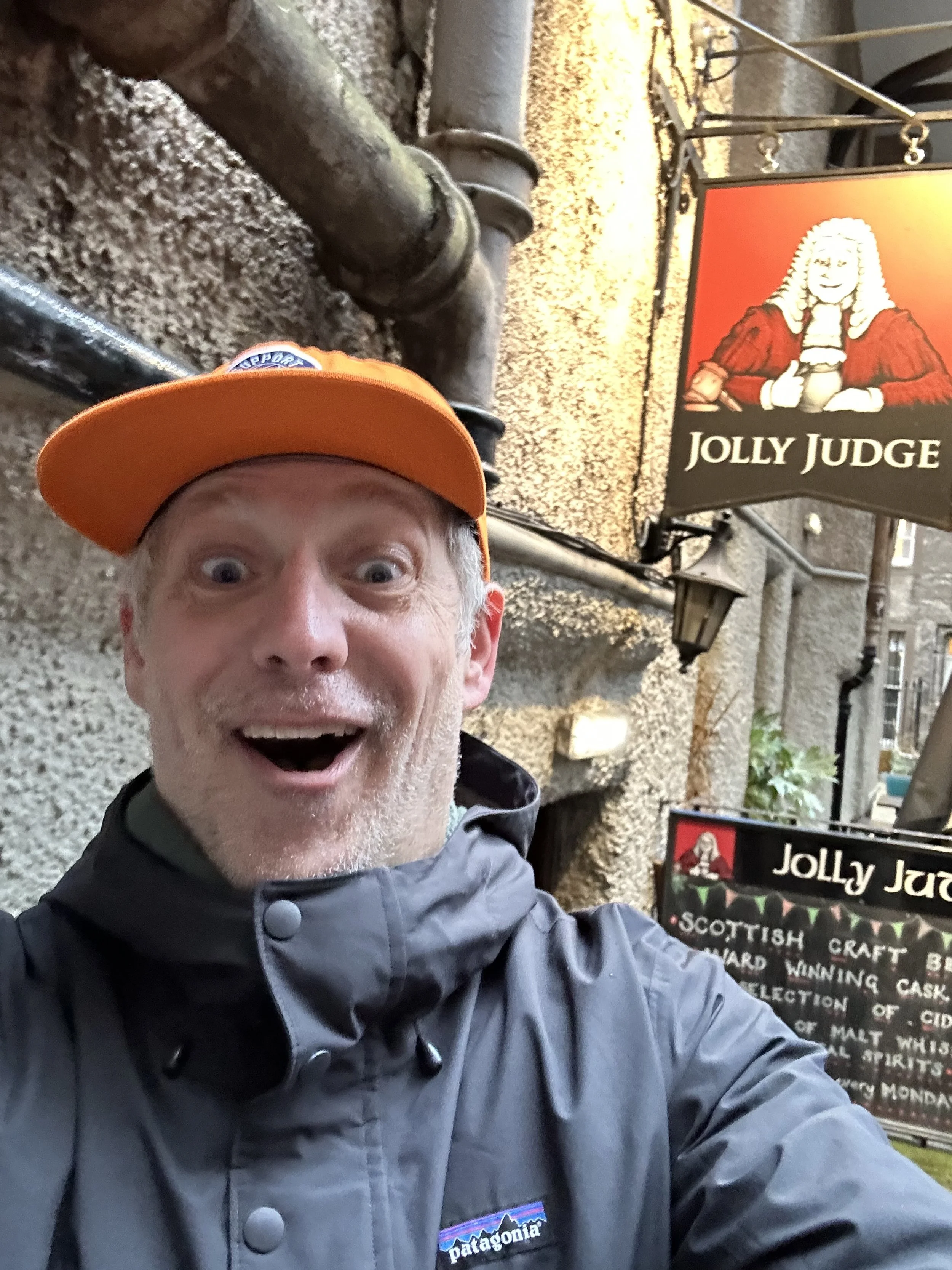 A cheerful man taking a selfie outside a pub with a sign that reads 'Jolly Judge'. The man is wearing an orange cap and a gray Patagonia jacket, standing against a stone wall with pipes and a lantern.
