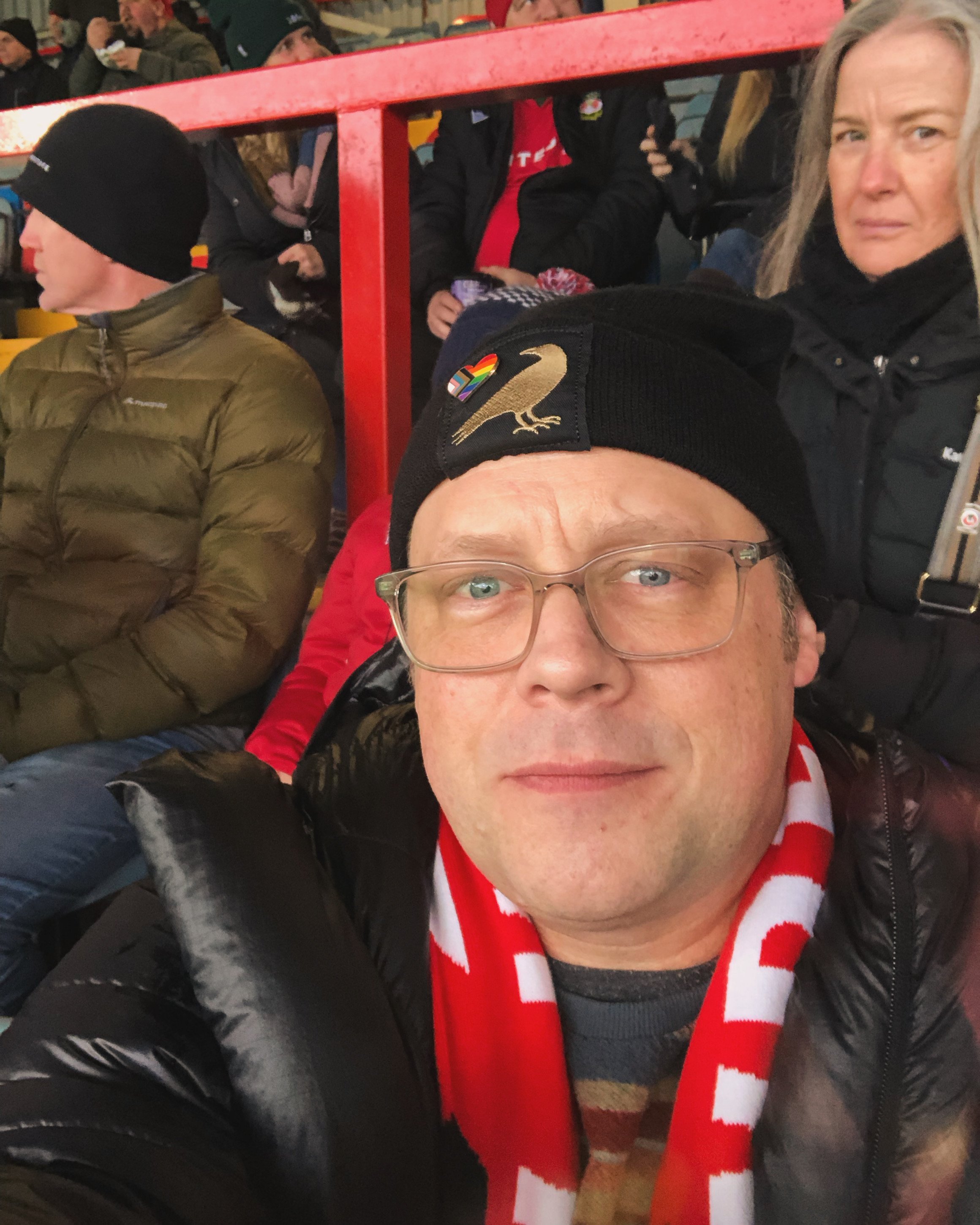 A man wearing glasses, a black beanie with a gold and rainbow heart pin and a bird patch, a red and white scarf, and a black jacket taking a selfie at a sporting event with other spectators in the background.