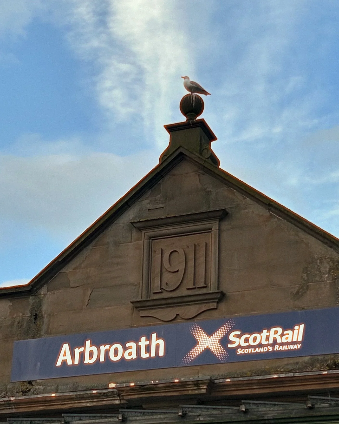 The entrance of a building with the year 1911 carved into the stone facade. On top of the building, there is a seagull perched on a decorative finial. Below the building facade, there is a digital sign displaying the text 'Arbroath' and 'ScotRail Sco