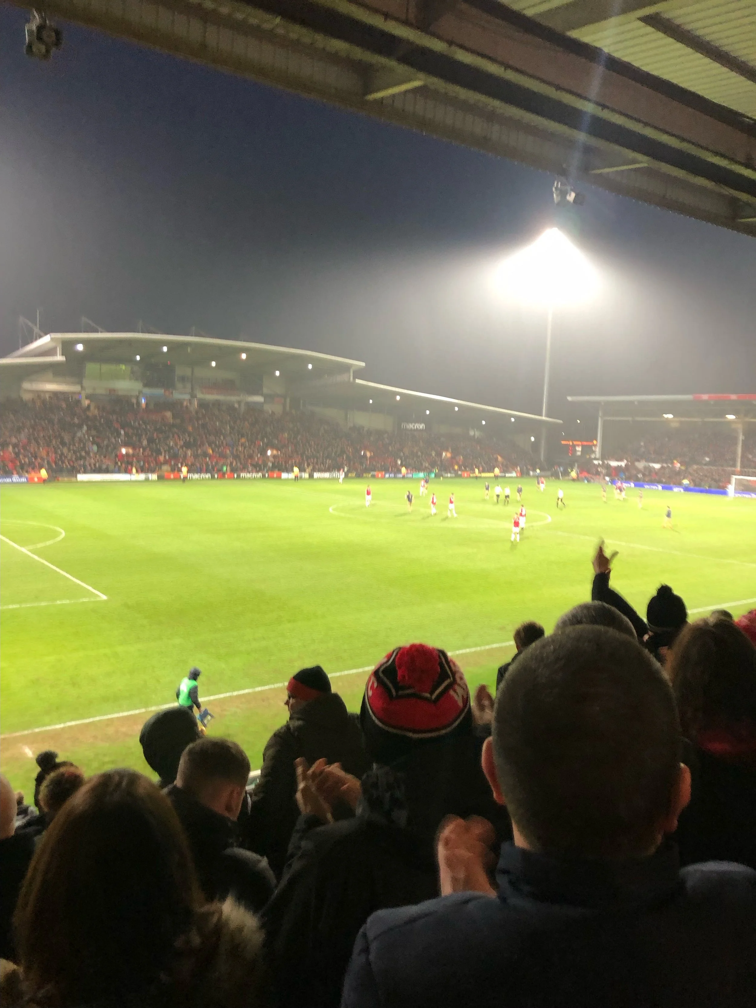 View of a soccer stadium filled with spectators at night, with players on the field and bright stadium lights overhead.