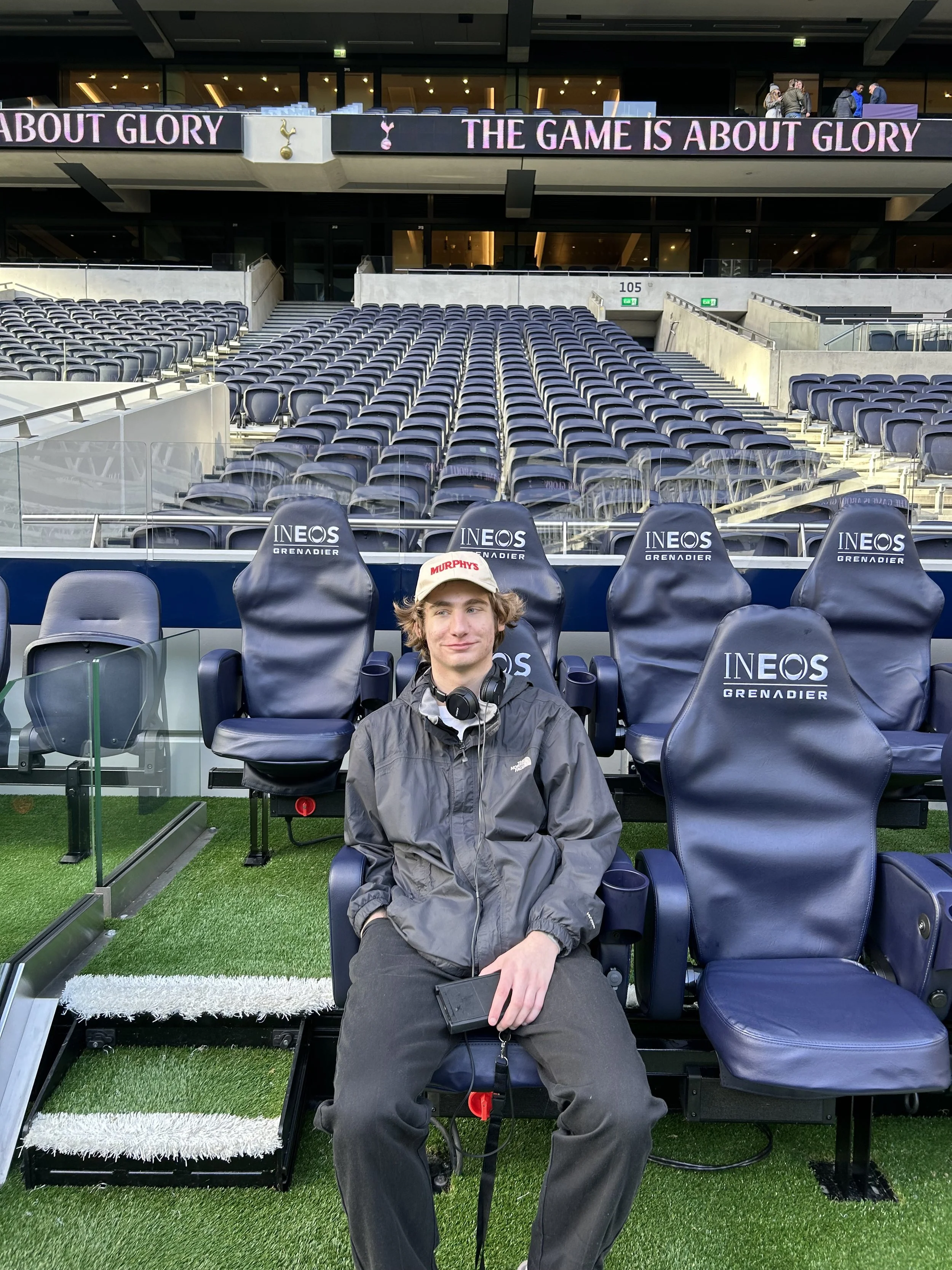 A young man sitting on a stadium seat in a football (soccer) stadium, with multiple empty seats and a large digital advertising banner in the background. The banner reads 'THE GAME IS ABOUT GLORY' and 'ABOUT GLORY'.