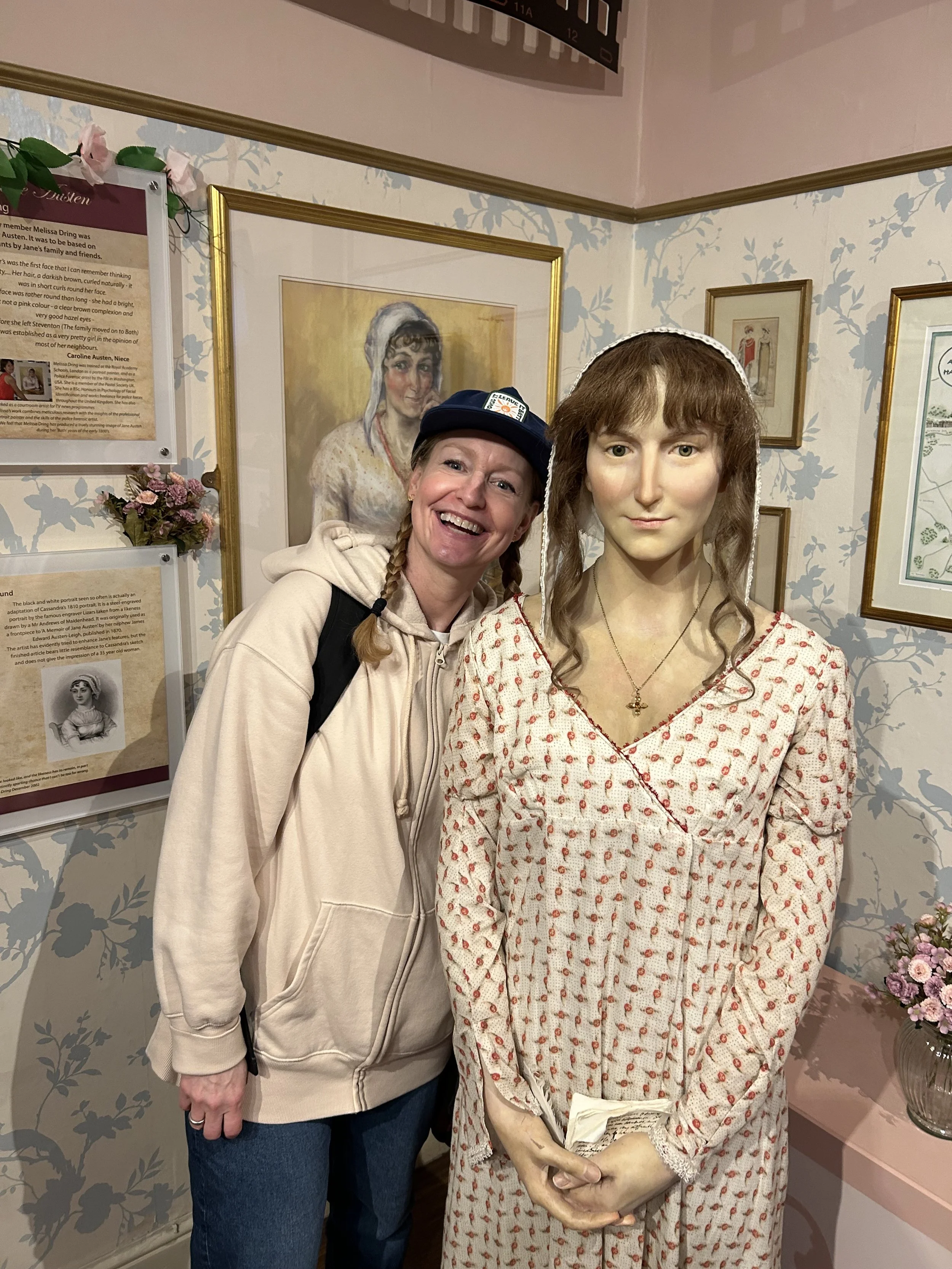 A woman smiling next to a life-sized wax figure of a woman in a patterned dress, holding a book in her hands, in a museum setting