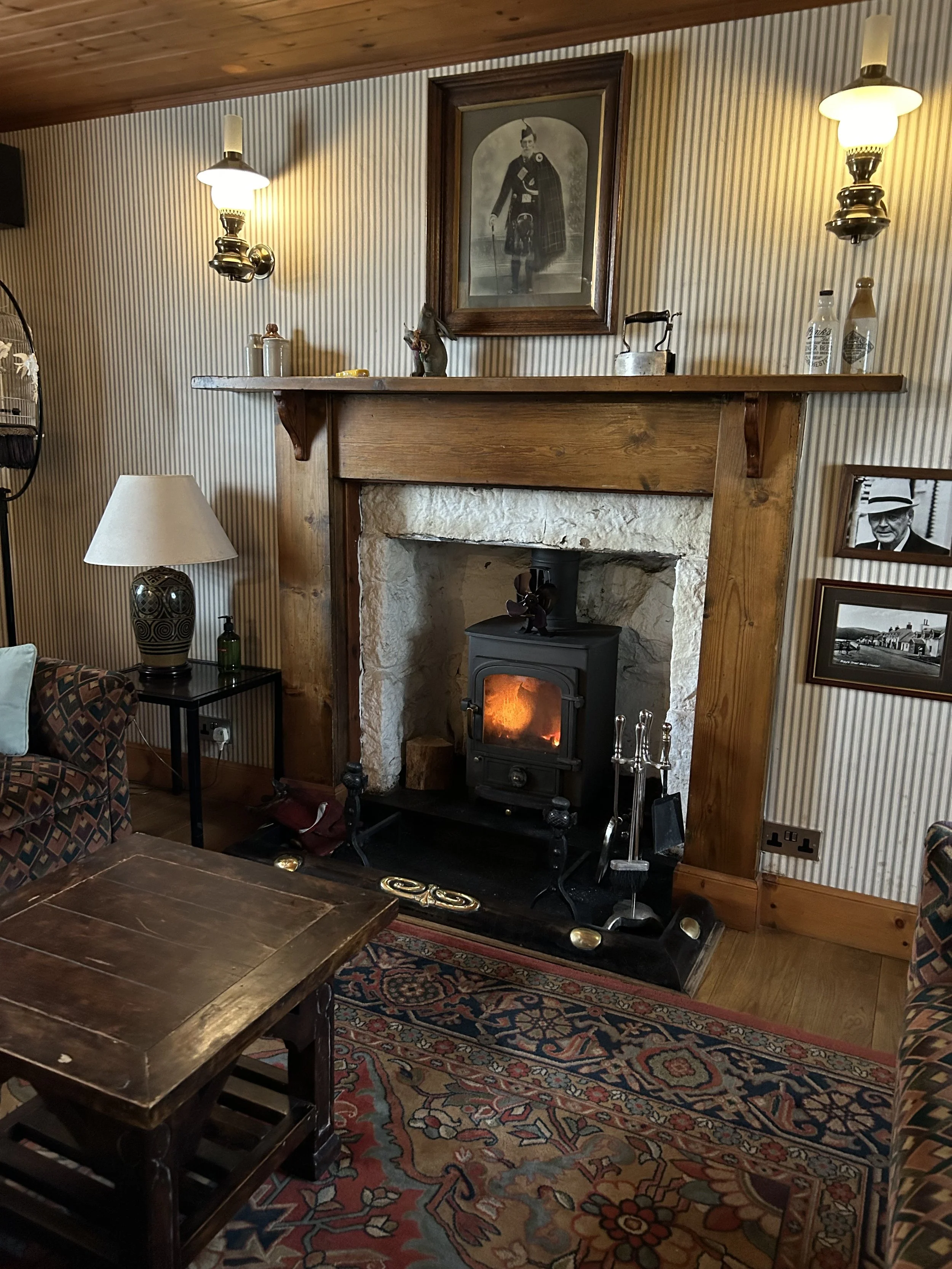 Cozy living room with a wood fireplace, framed black-and-white portrait, wall-mounted lamps, patterned rug, wooden furniture, and decorative items.