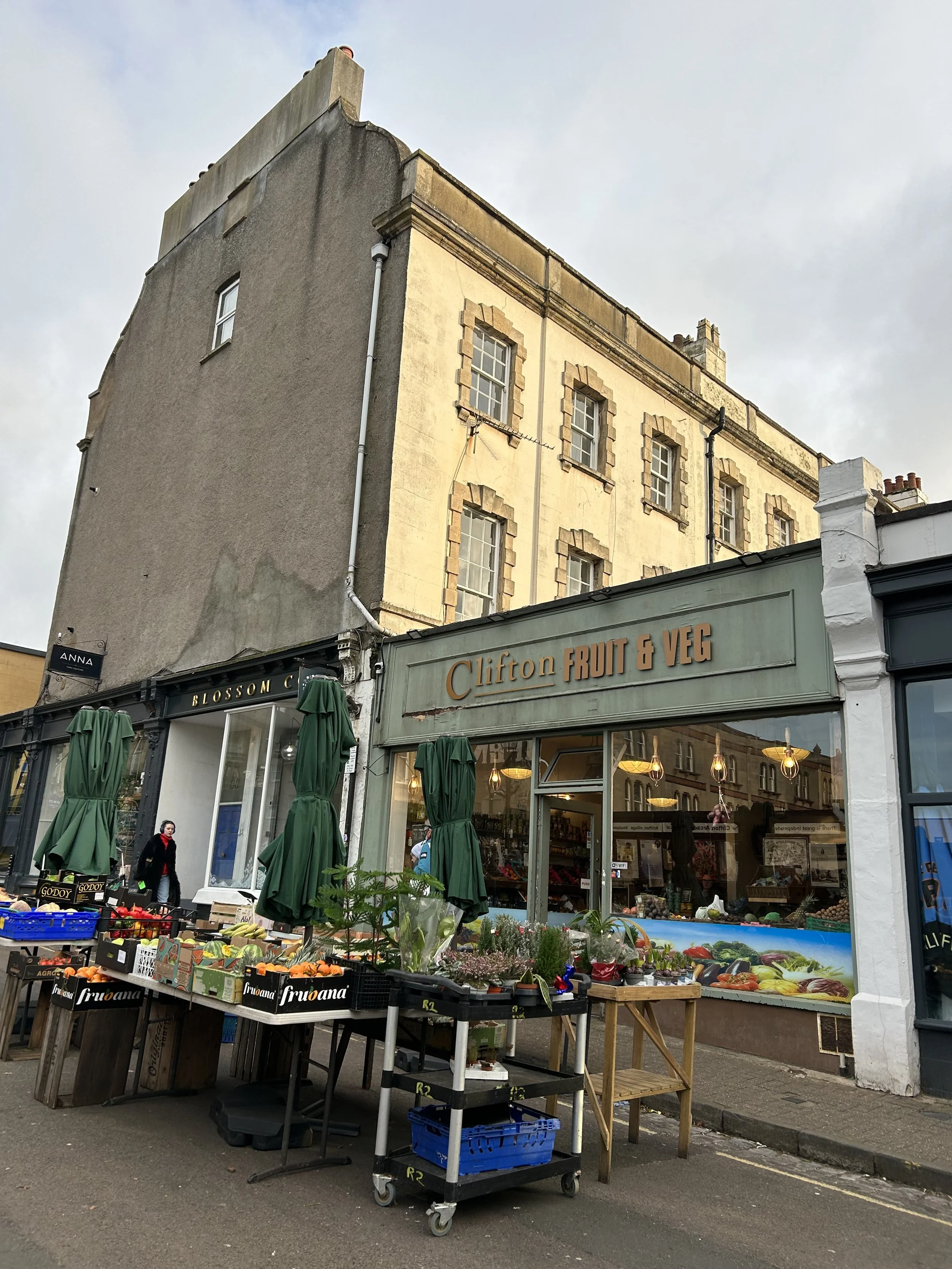 Street view featuring a fruit and vegetable shop named 'Clifton Fruit & Veg' with outdoor produce displays, including crates of fruit and vegetables, and green umbrellas, in front of an old multi-story building.