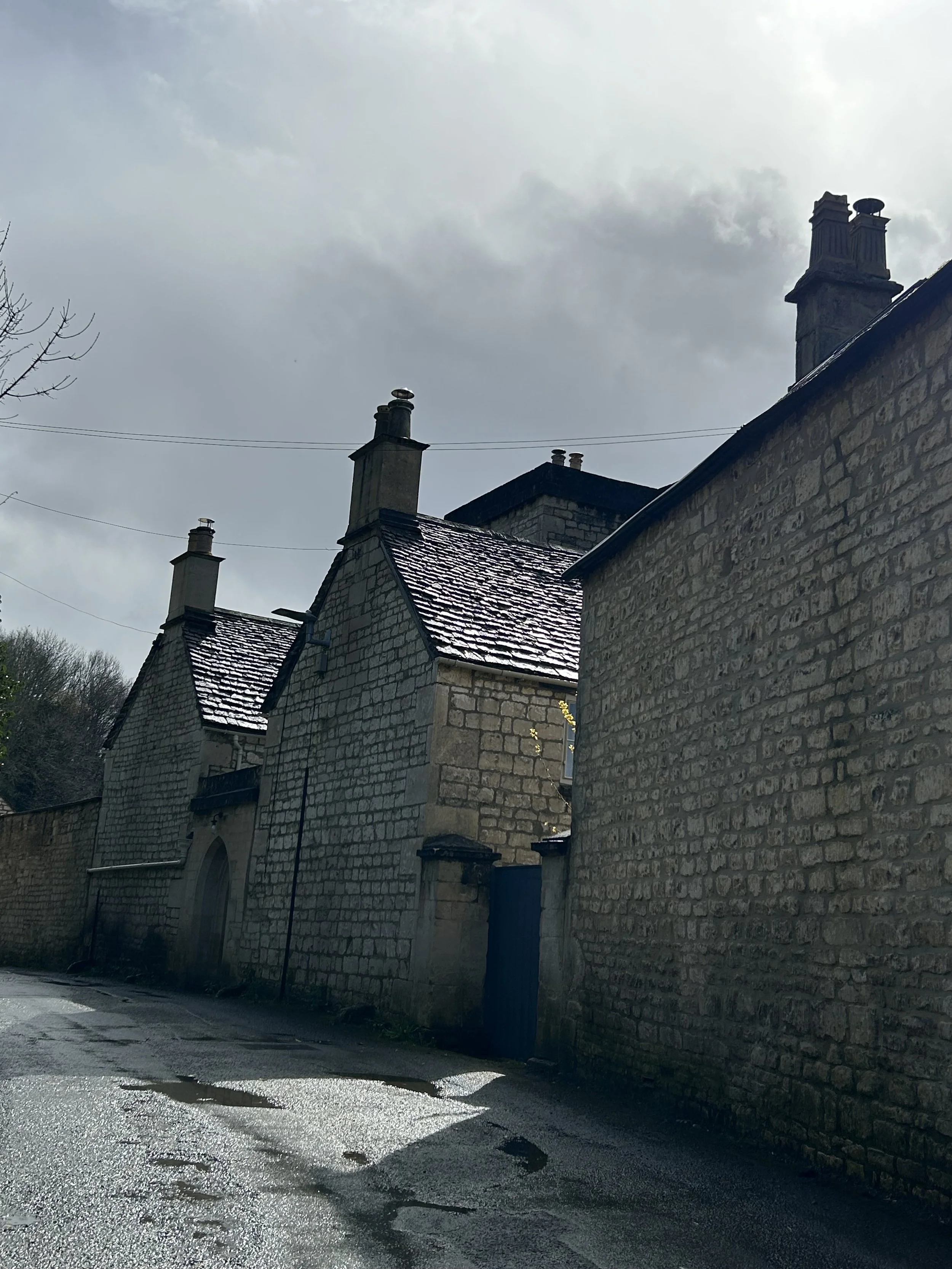 Old stone buildings with chimneys and tiled roofs on a rainy street under cloudy sky.