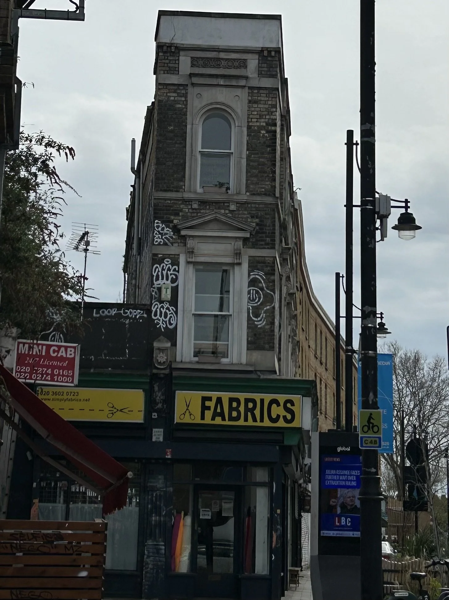 A multi-story building with a fabric store on the ground floor, featuring a yellow sign that reads 'Fabrics' with scissors icon. The upper floors have brick and stone architecture with ornate window details, graffiti on the wall, and a street lamp an