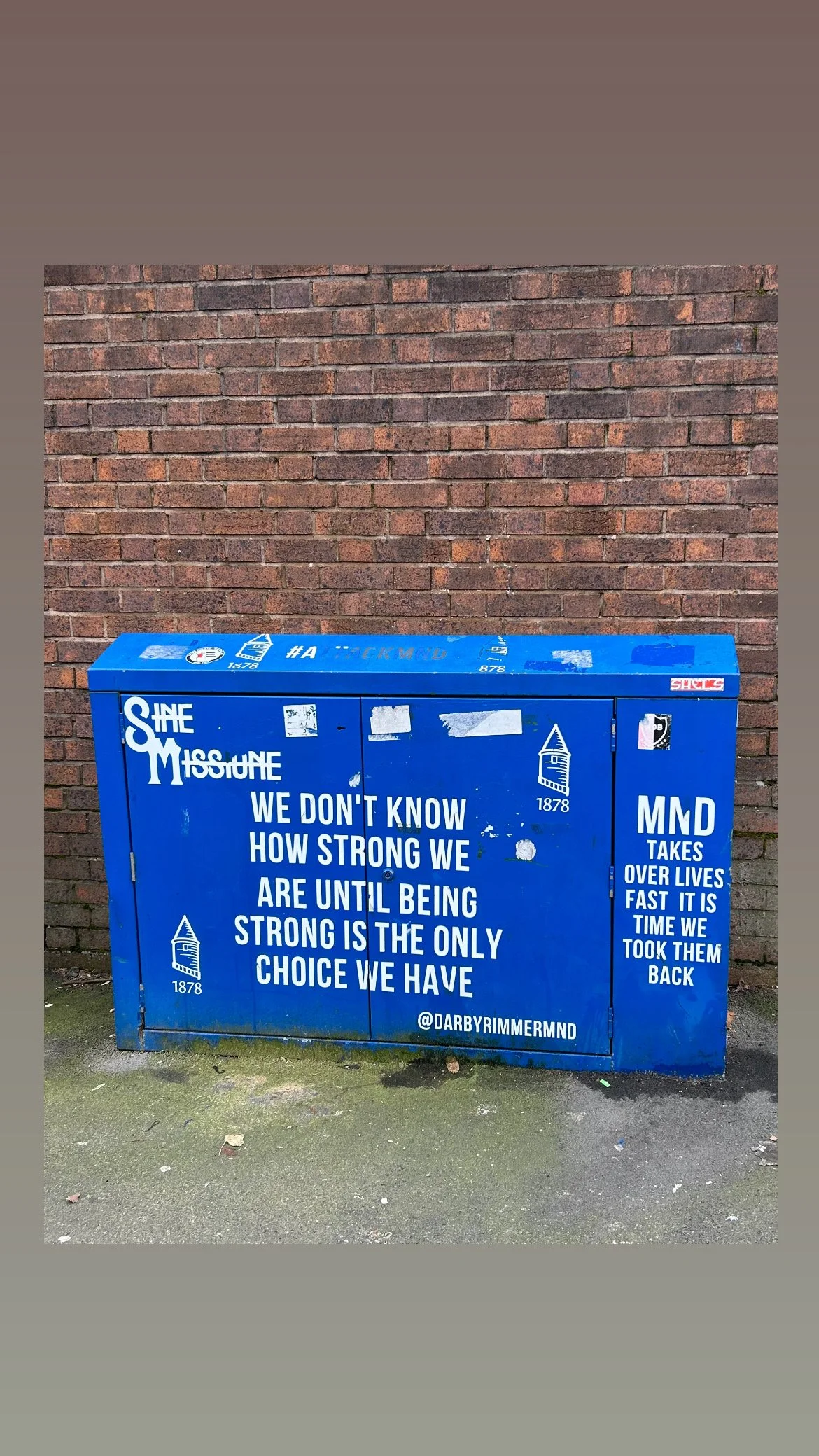 Blue electrical box with white text and Facebook handle, placed in front of a brick wall and on a concrete sidewalk.