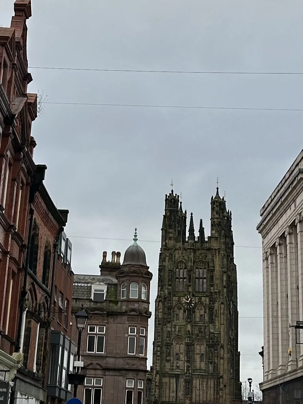 A city street scene with historic buildings and a prominent dark stone church tower under an overcast sky.