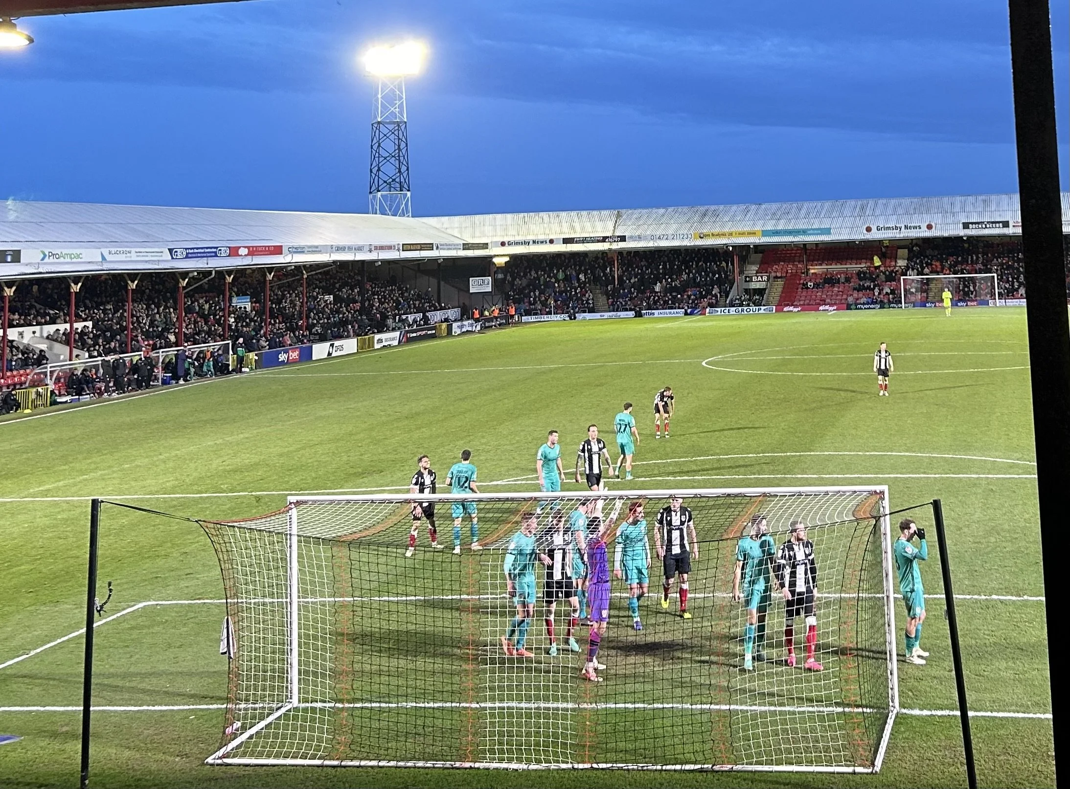 Soccer players on the field near the goal post, with a crowd of spectators in the stands under a evening sky.