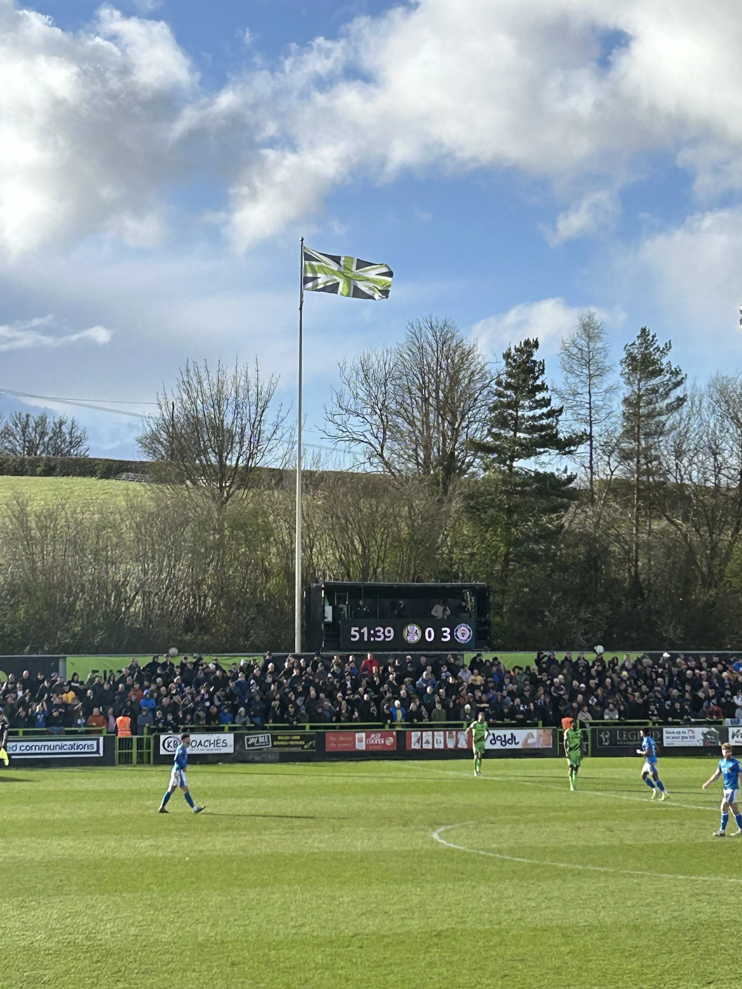 A soccer match at a stadium with a score of 0-3, a time of 51:39 on the scoreboard, a large crowd of spectators in the stands, and a British flag flying on a pole above the field, with a backdrop of trees and partly cloudy sky.