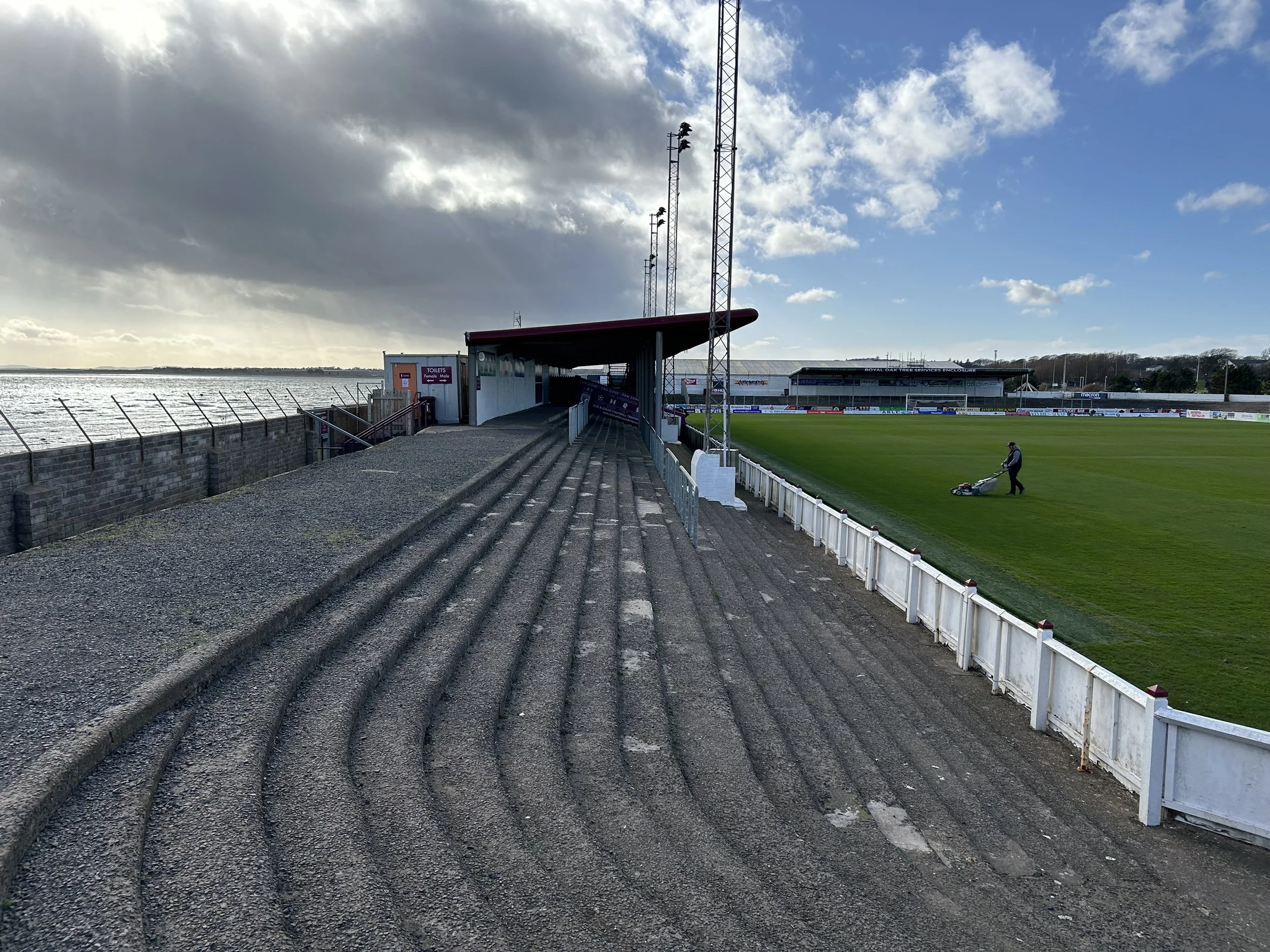 Empty sports stadium with a person mowing the grass on the field, sky with clouds and sunlight coming through.