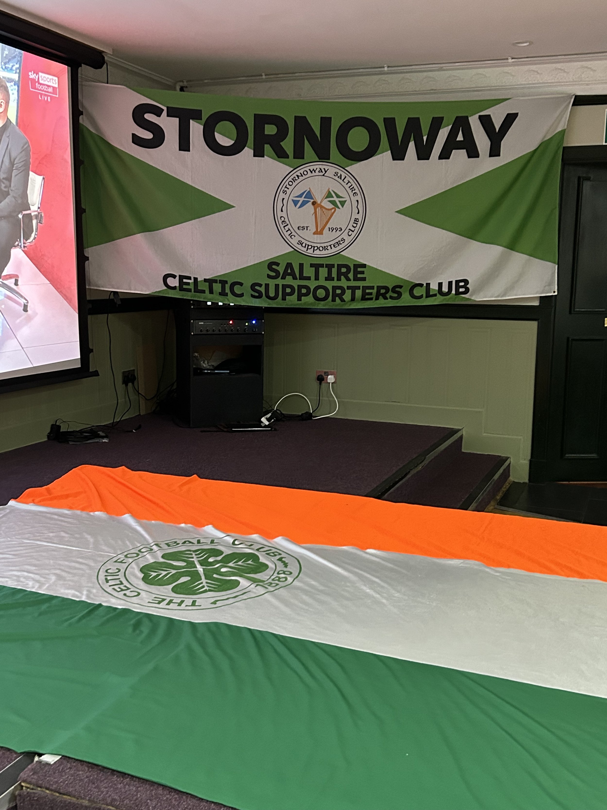 Banner for Stornoway Saltire Celtic Supporters Club hanging on a wall in a room with a stage, alongside a Celtic flag with orange, white, and green colors laid out on the floor, and a projector screen showing a person in a suit.