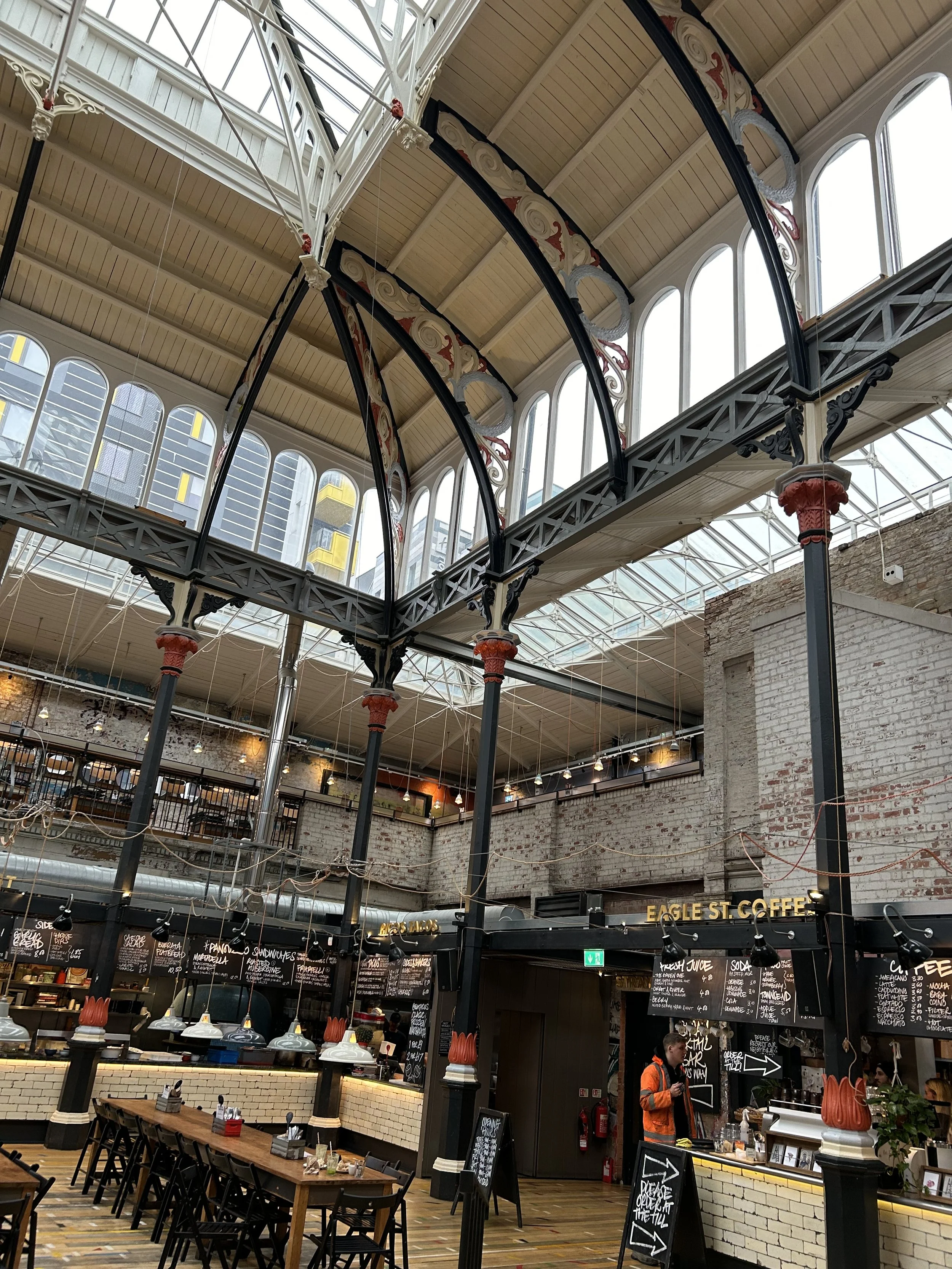 Interior of a modern, industrial-style café with exposed brick walls, high arched windows, and decorative iron and wood architectural elements.