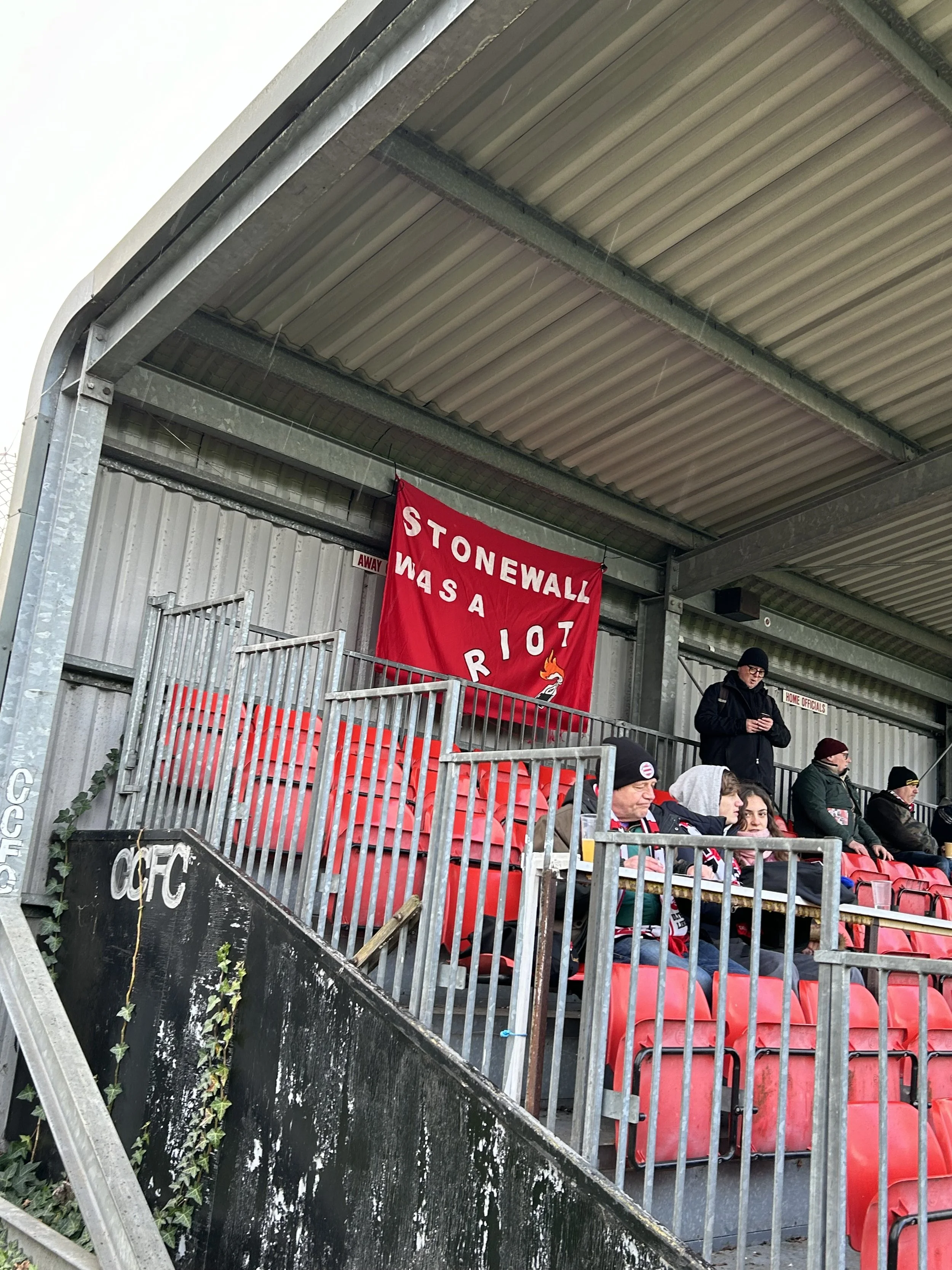 Spectators sitting on red seats at a sports stadium, with a red banner that reads "Stoneywall Was a Riot" hanging behind them. Some people are standing, dressed in winter clothing.