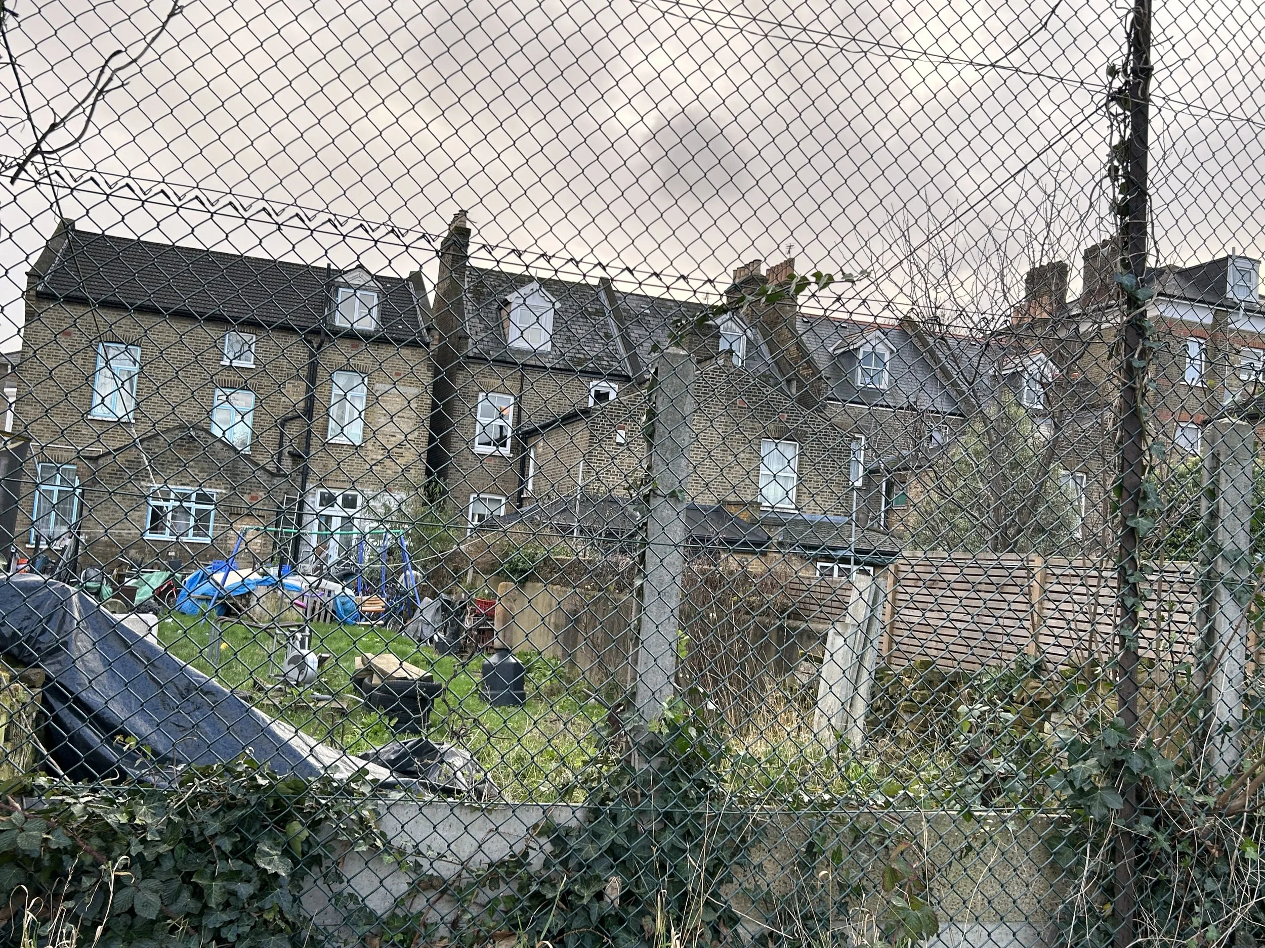 Residential houses viewed through a chain-link fence, with a backyard containing gardening tools, bicycles, and a small grassy area.