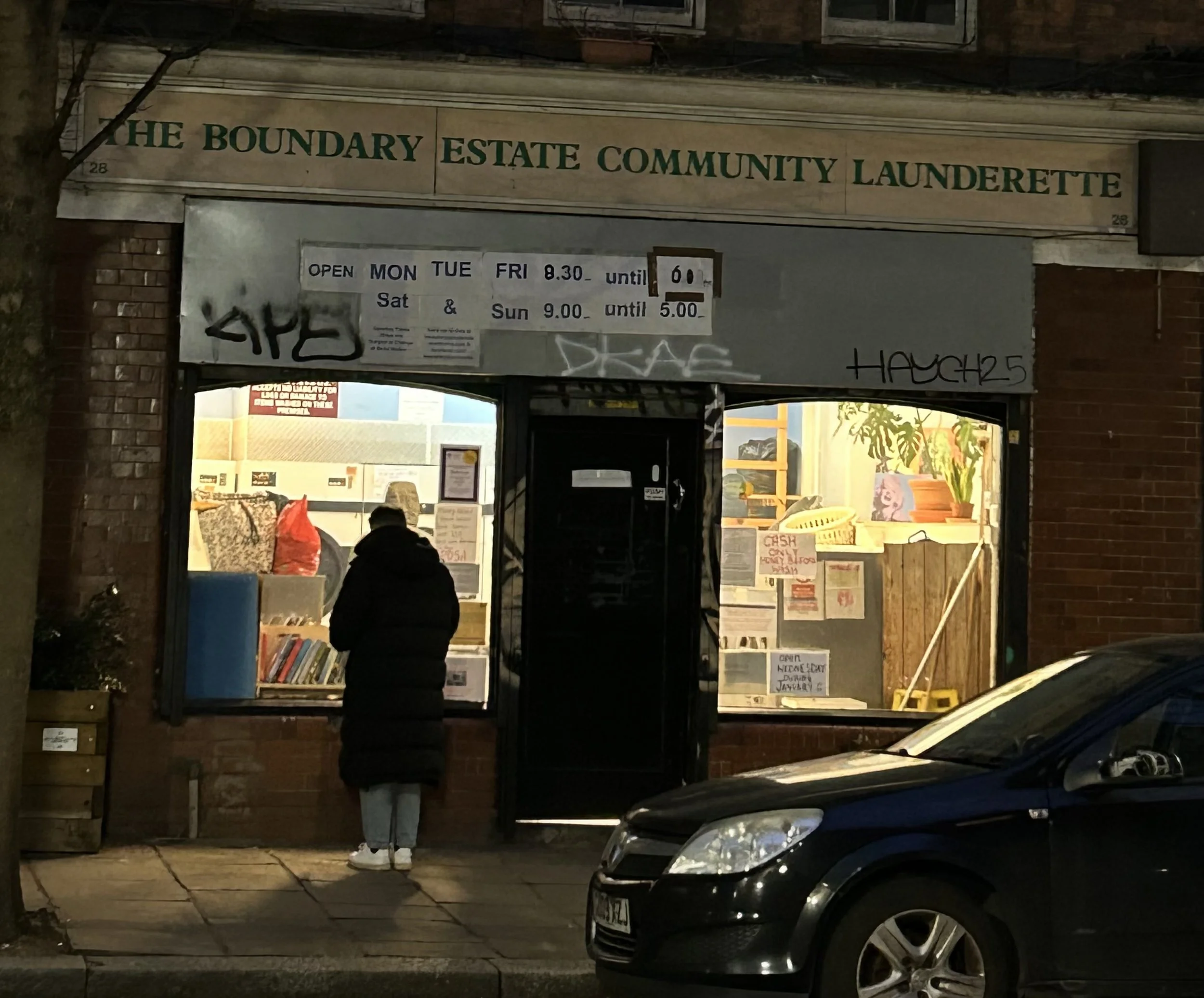The Boundary Estate Community Laundrette storefront at night, with a person in a dark coat and light pants standing outside looking in. There are two large windows displaying laundry items and a potted plant inside, and a black car parked in front.