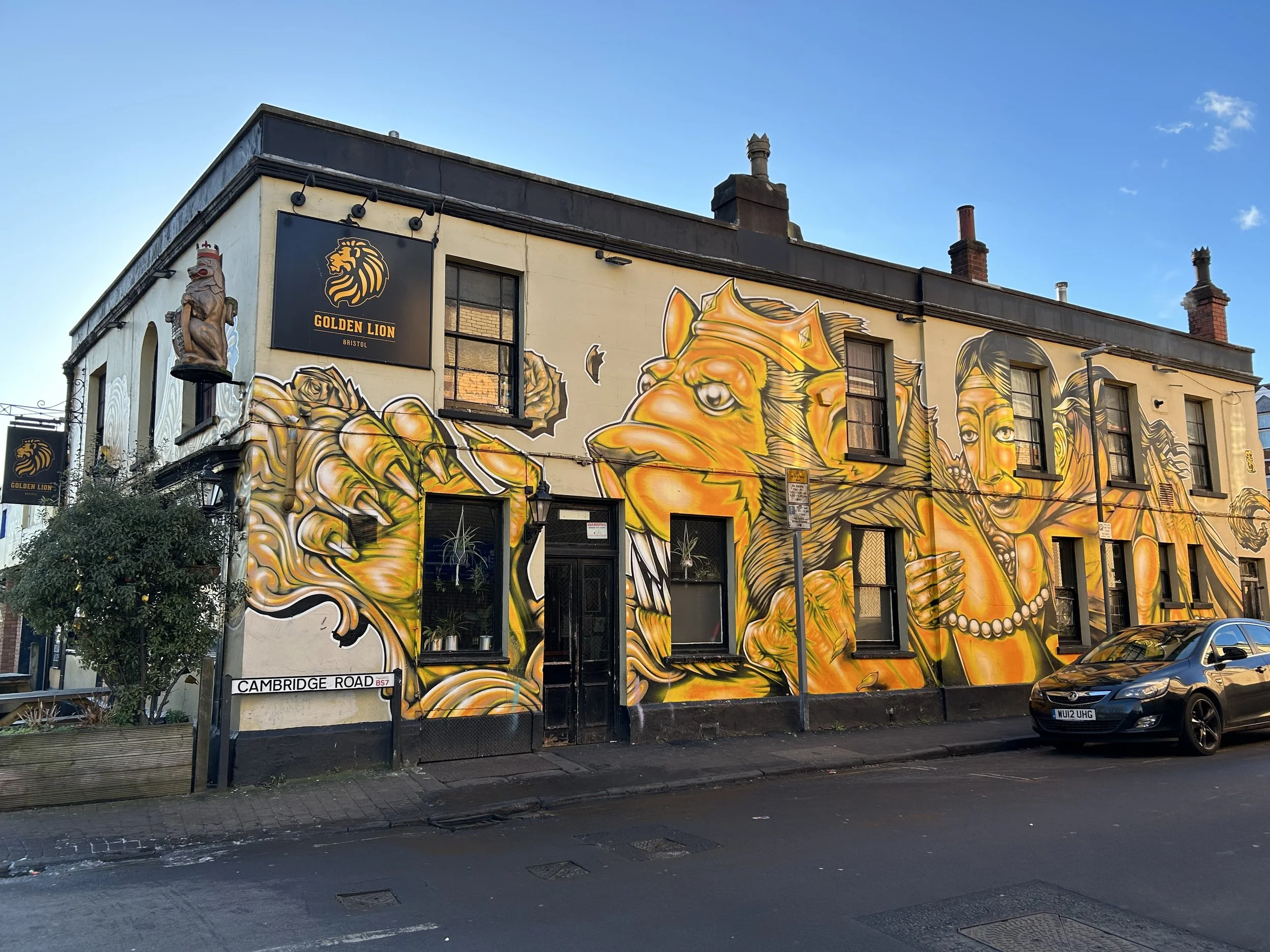 A building with colorful graffiti art featuring a lion, a woman with beads, and a hand, with the sign 'Golden Lion' on the corner of Cambridge Road in Bristol.