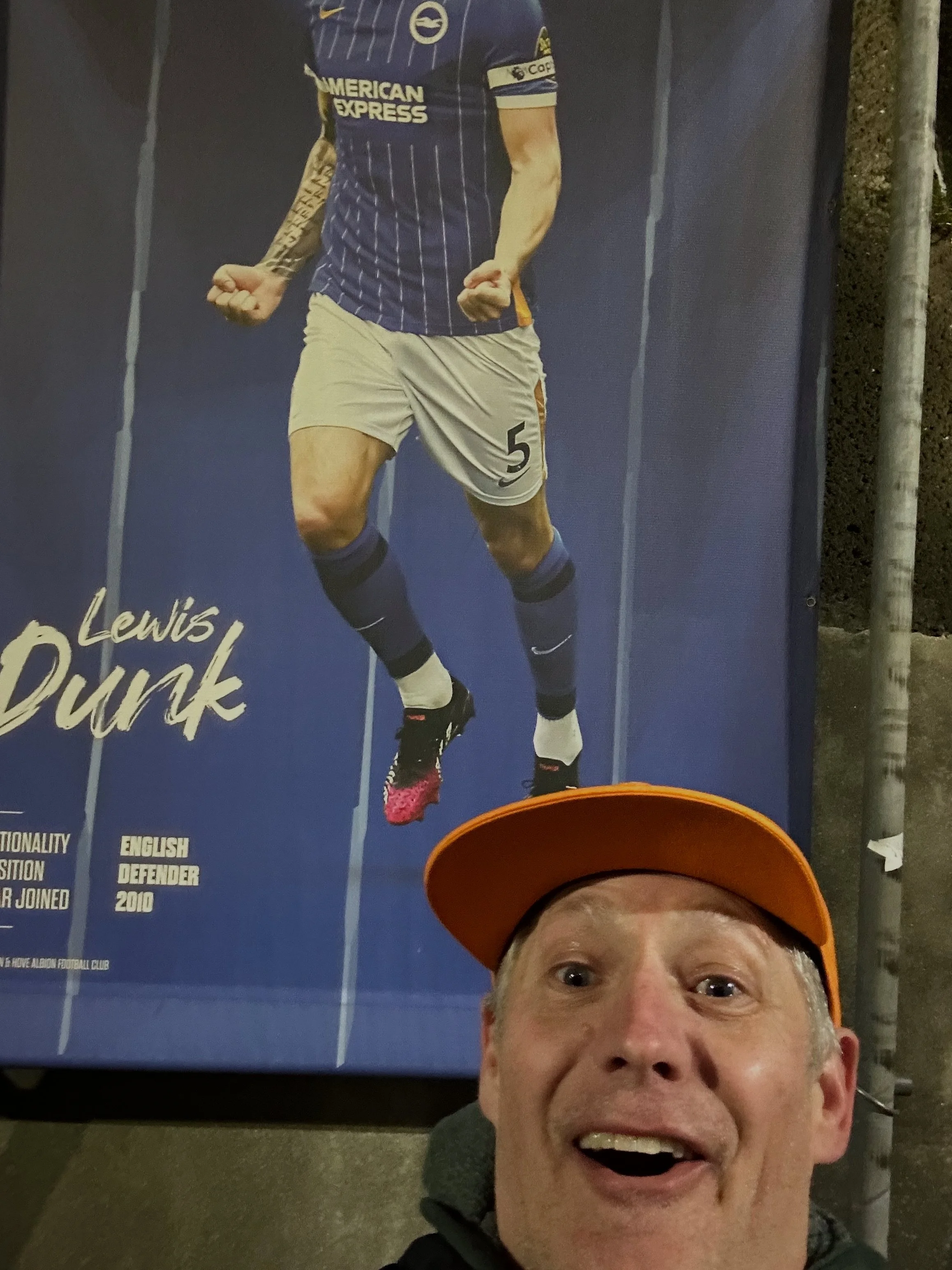 Author, Todd Sith wearing an orange cap smiling in front of a poster of soccer player Lewis Dunk celebrating, wearing a blue and white jersey and shorts, with the number 5.