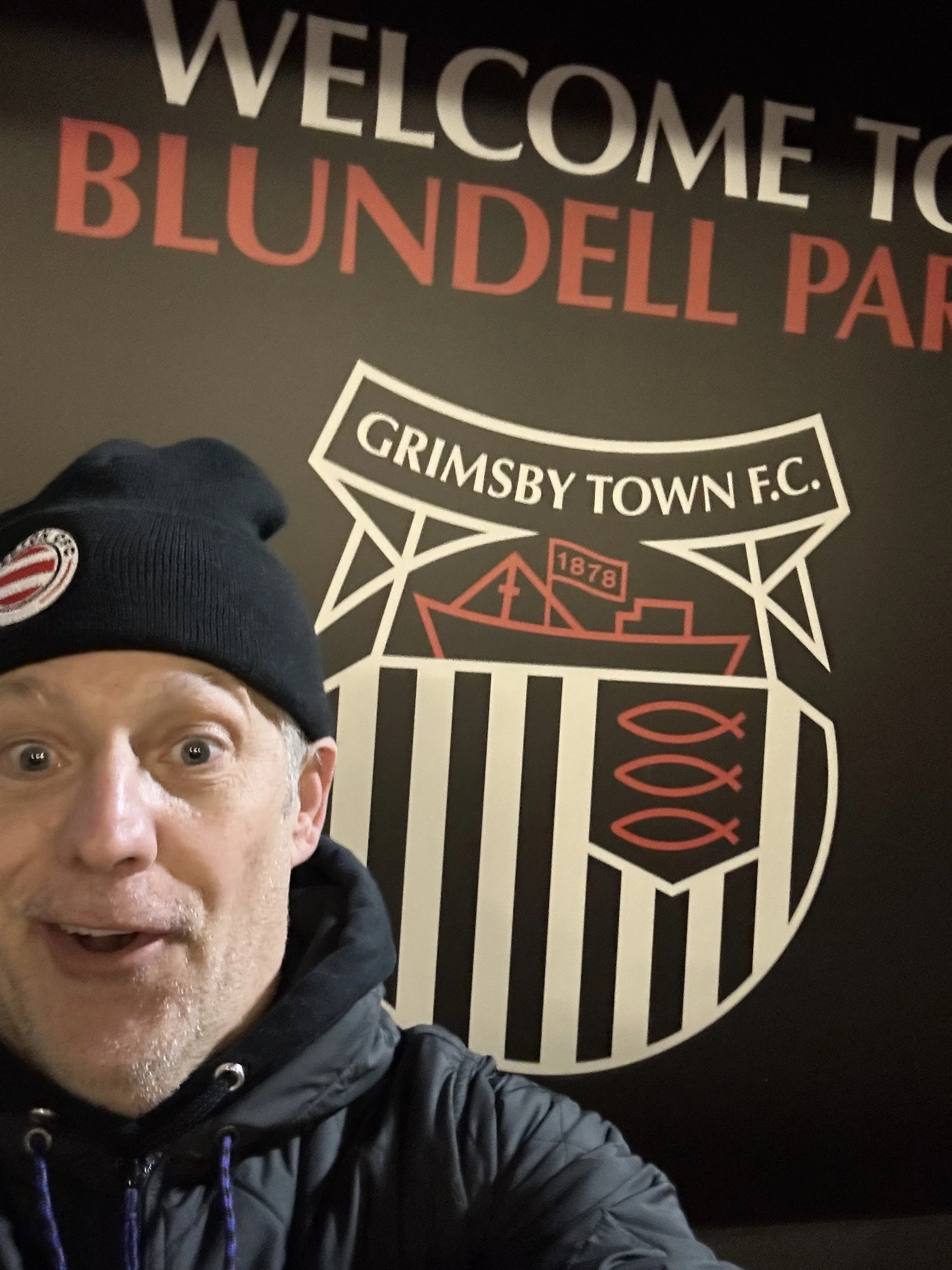 Author Todd Smith taking a selfie in front of a wall with the Grimsby Town F.C. logo and a sign that reads "Welcome to Blundell Park".