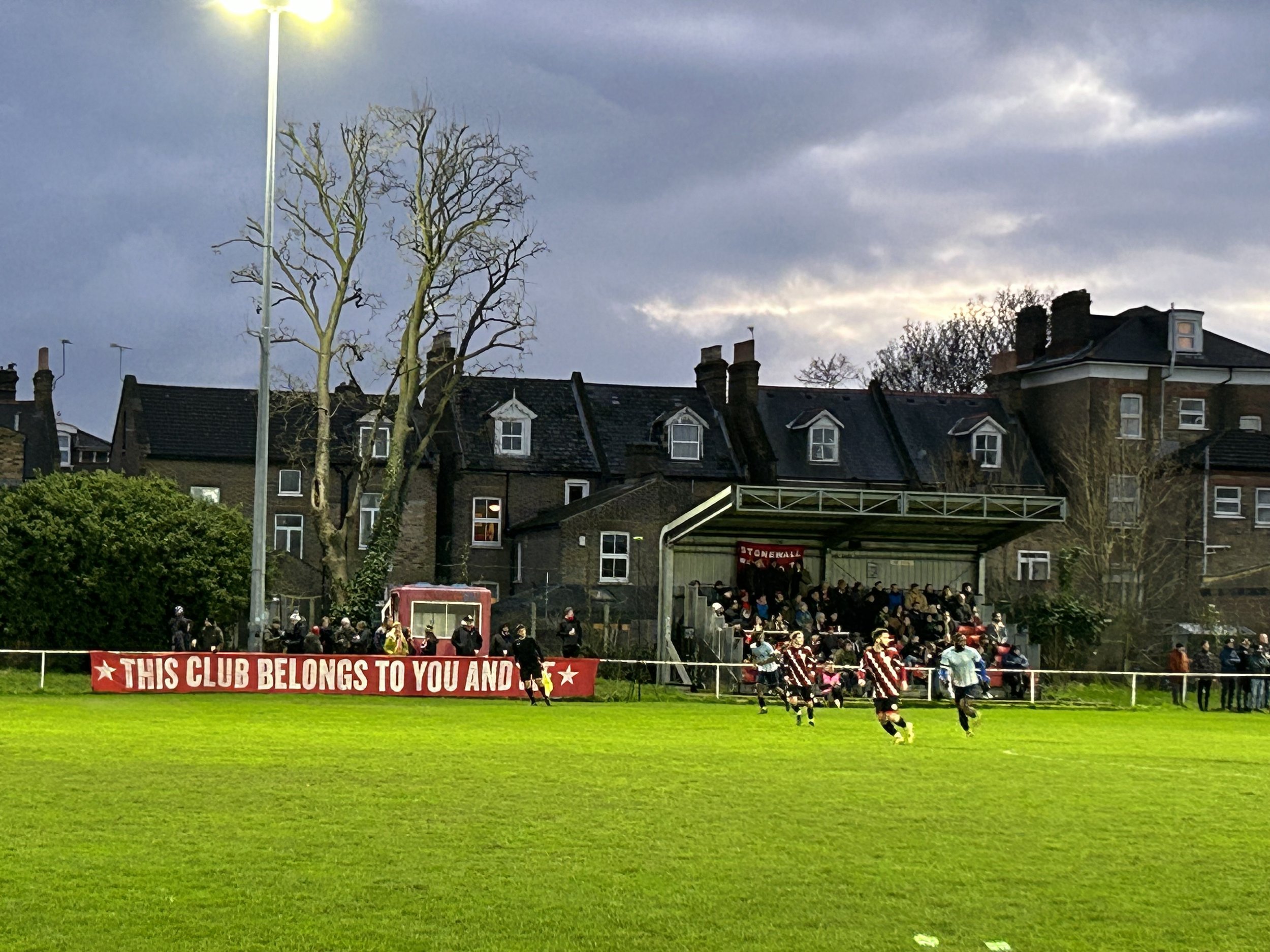 A soccer game taking place in a stadium with green grass, spectators on the stands, and a row of houses and leafless trees in the background. The sky is overcast, and there is a red banner on the fence that says, 'THIS CLUB BELONGS TO YOU AND.'