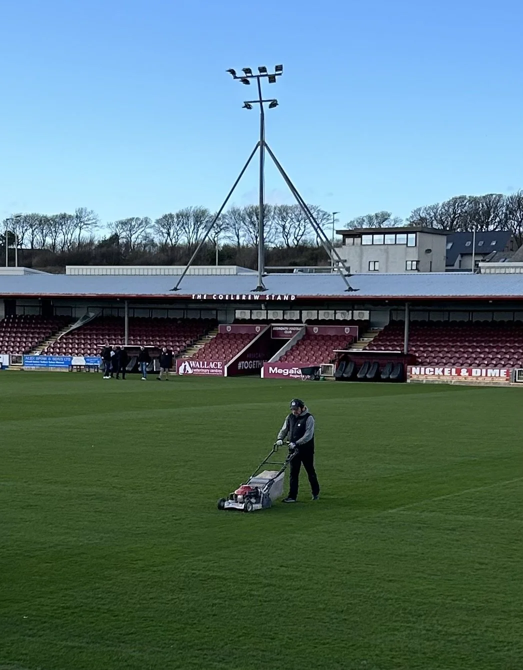 A person mowing the grass on a sports field, with empty stadium seats and a tall spotlight in the background.