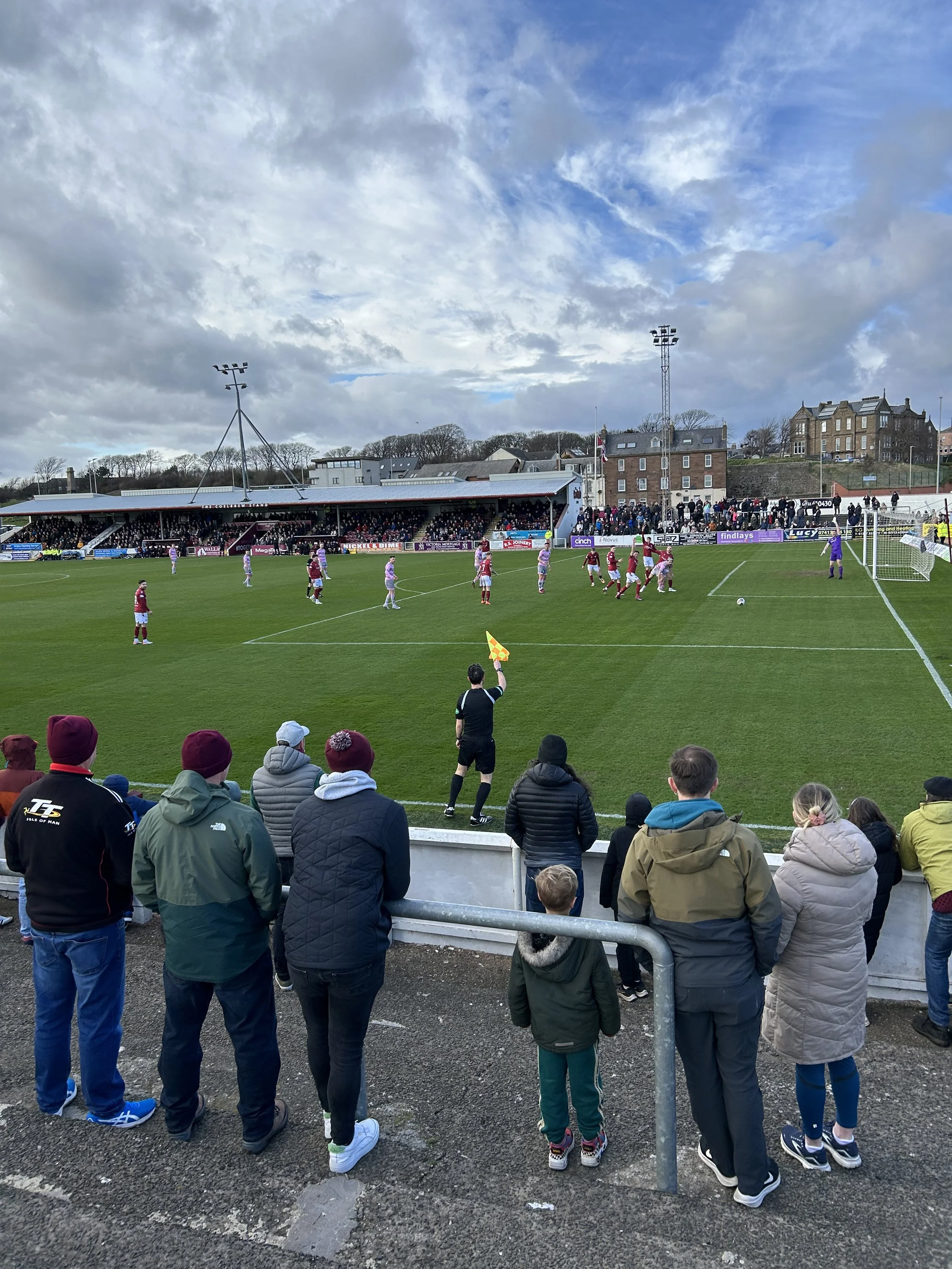 A soccer match is taking place at a stadium with spectators watching from the stands and sidelines. Players are on the field near the goal, with the referee holding a flag.