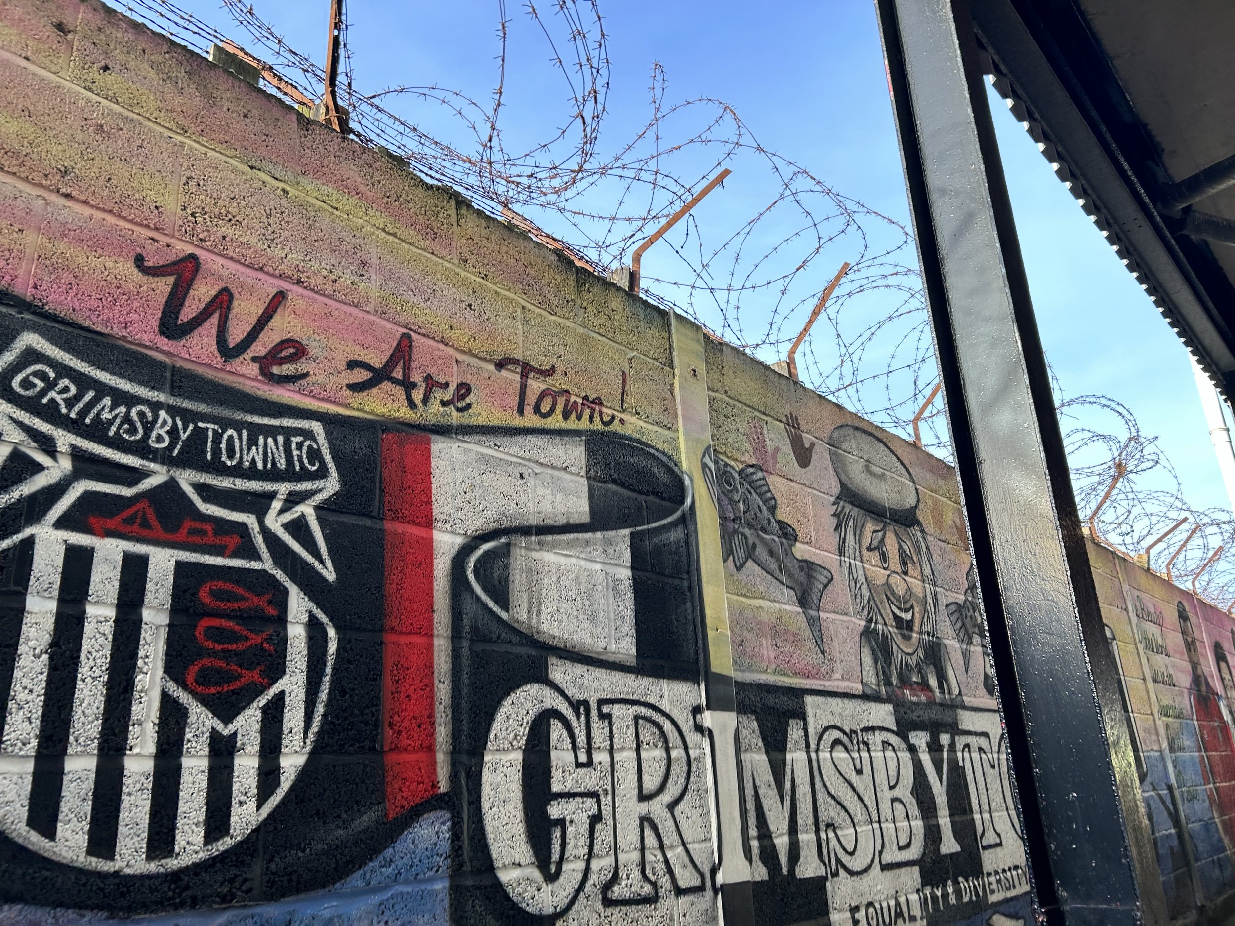 Graffiti art on a brick wall featuring the Grimmsby Town FC emblem, a cartoon character with long hair, and the phrase 'We Are Town!' with barbed wire on top of the wall.