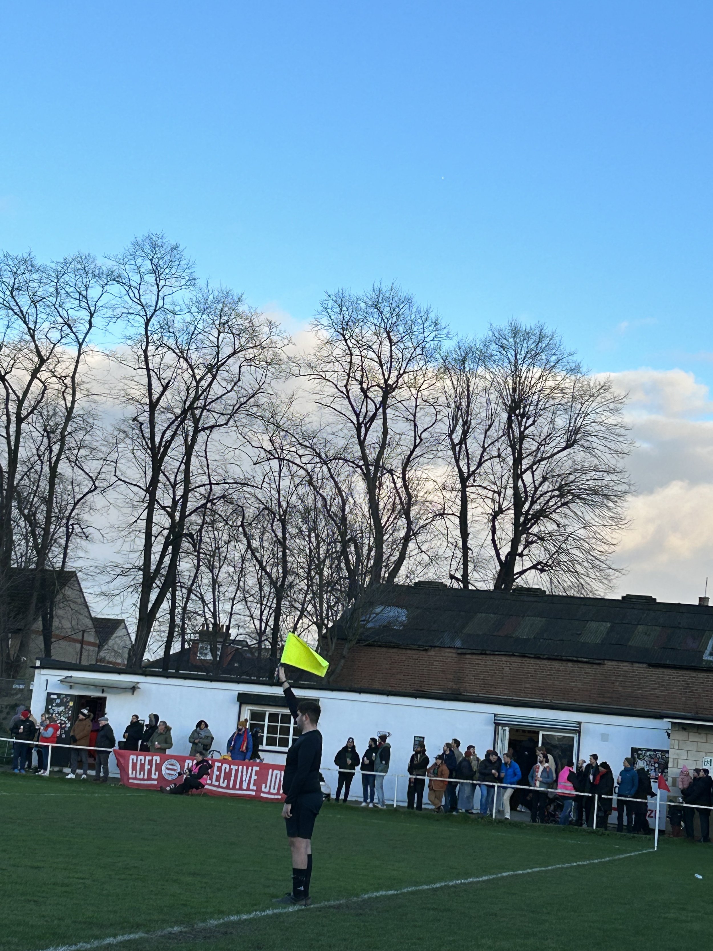 A soccer referee standing on the field holding a yellow flag during a game while spectators watch behind a barrier, with leafless trees and a building in the background under a blue sky with some clouds.