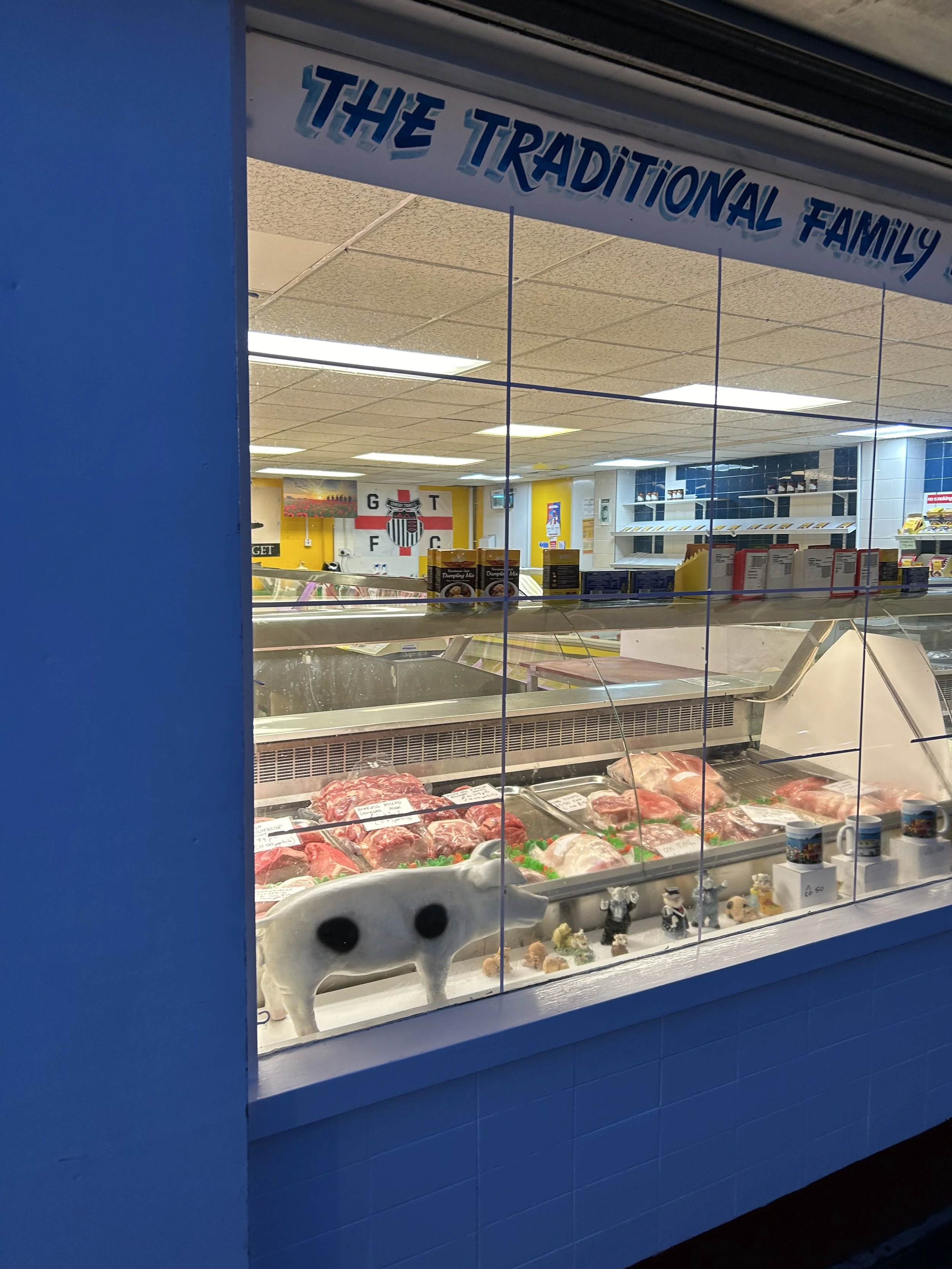 View of a butcher shop window displaying various cuts of raw meat, a pig-shaped plush toy, and small figurines on a white surface inside the store with a sign that reads 'The Traditional Family' above.