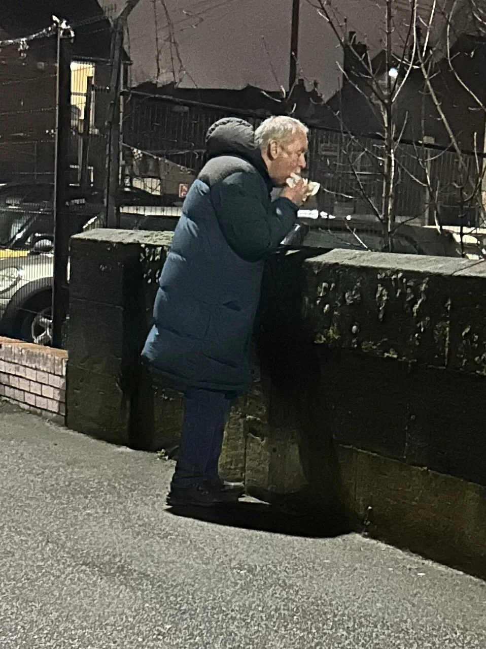 A man with gray hair wearing a blue puffer jacket standing on a sidewalk, drinking from a cup, near a brick and concrete wall, at night.