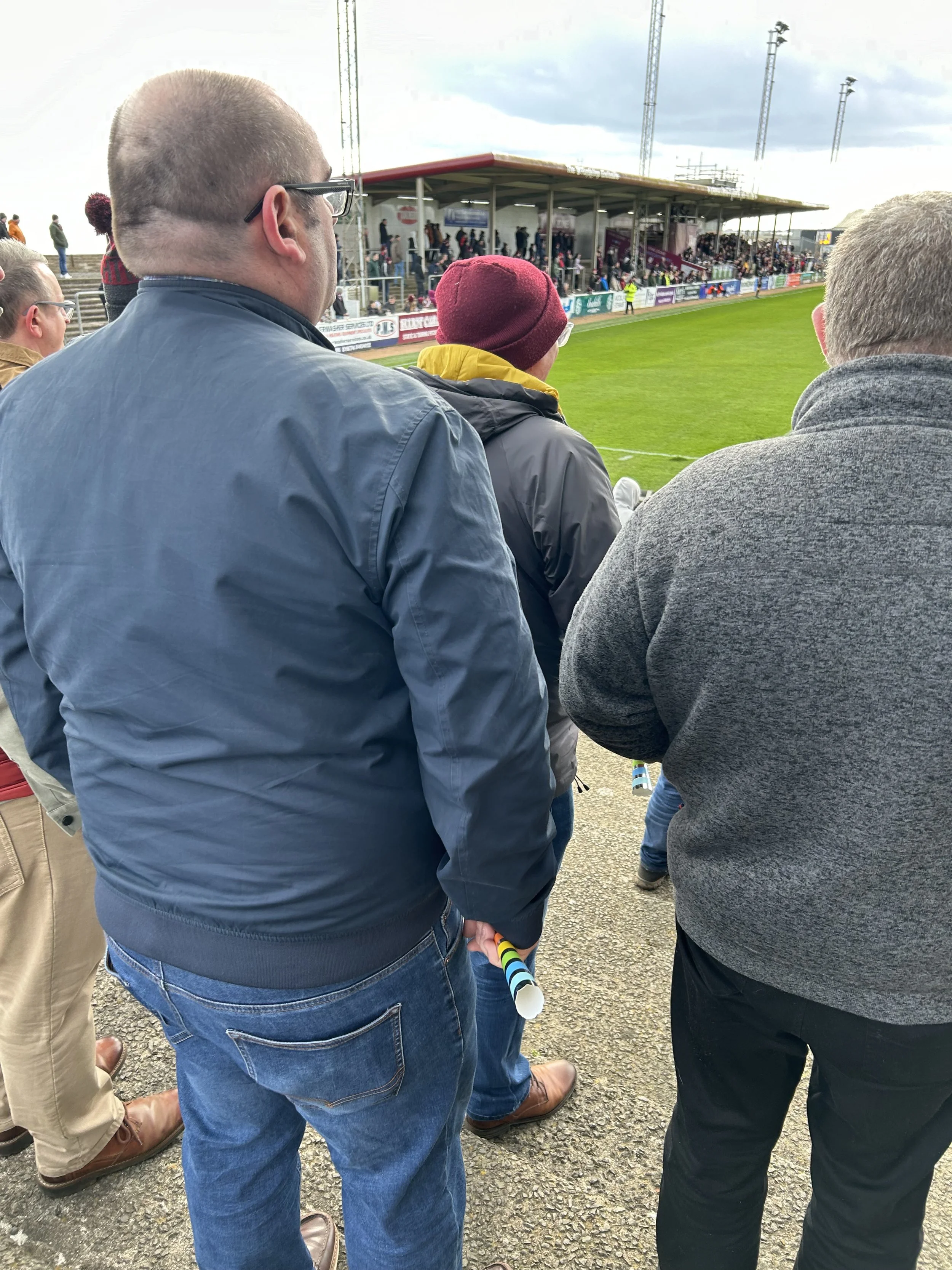People standing at the edge of a sports field watching a game, with stadium seating and spectators in the background.