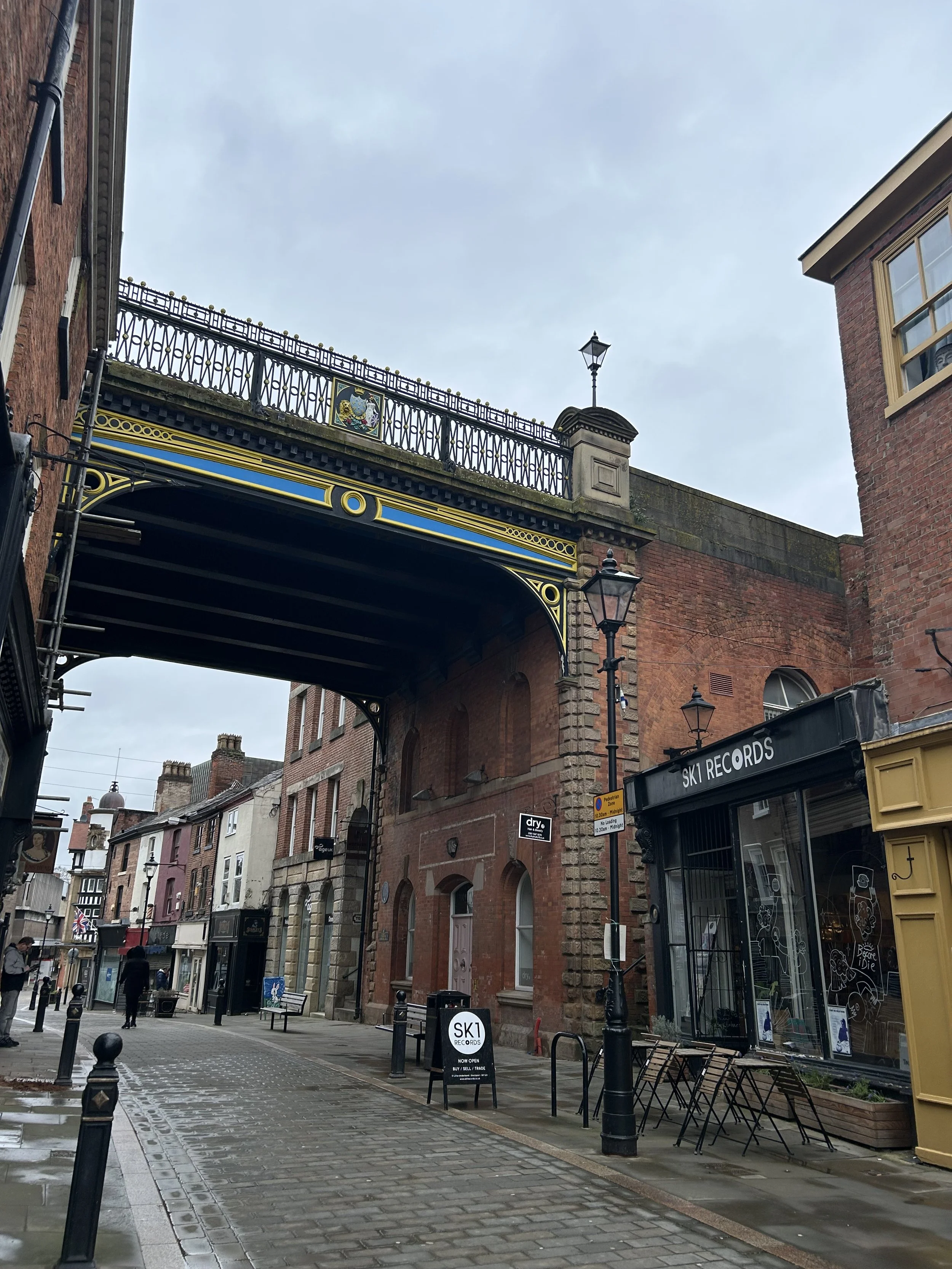 A street scene with brick buildings, a bridge overhead, and shops including Ski Records, on a cloudy day.