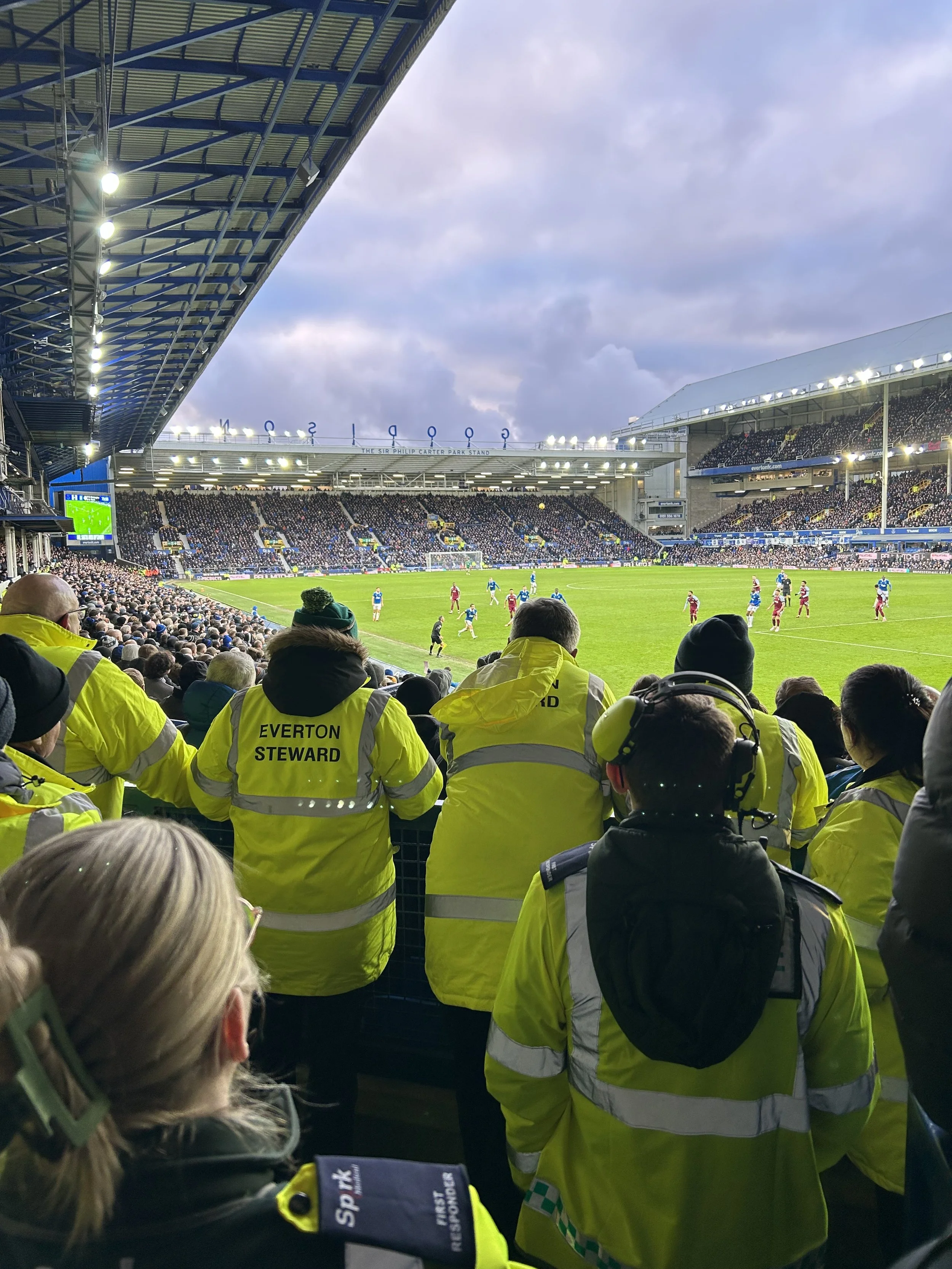 Spectators in high-visibility jackets at a soccer match watching the game on the field in a large stadium with a partially cloudy sky.