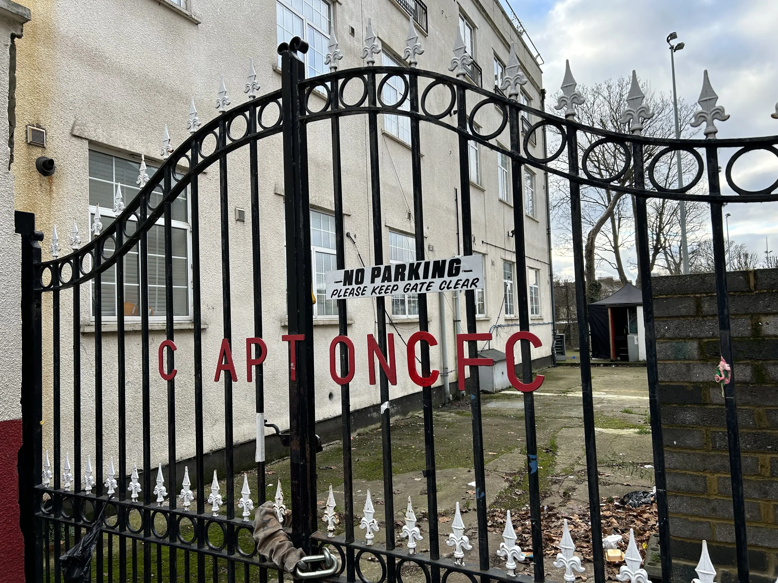 A black wrought iron gate with white decorative spikes at the top, attached to a brick wall on one side and a beige building on the other. The gate has red letters spelling, 'CAPT ON FC', with a sign that says, 'NO PARKING - PLEASE KEEP GATE CLEAR' h