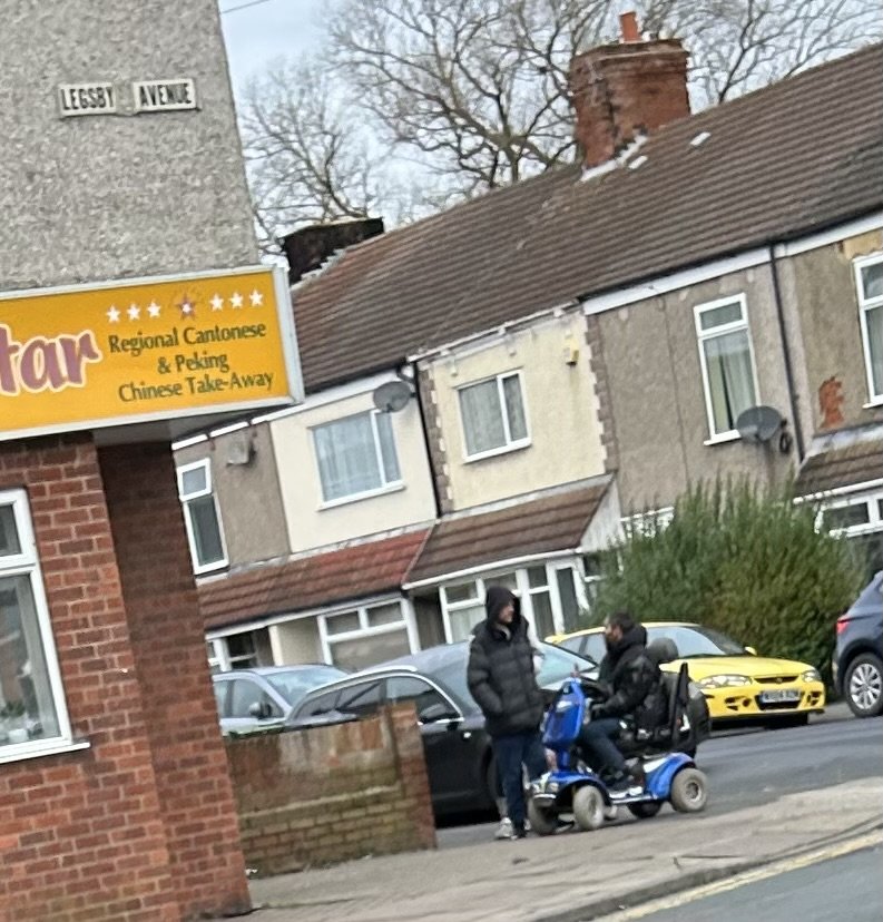 Two men are on a blue mobility scooter parked on the sidewalk in front of residential houses. One man is sitting on the scooter, and the other is standing beside him. There are parked cars behind them on the street.