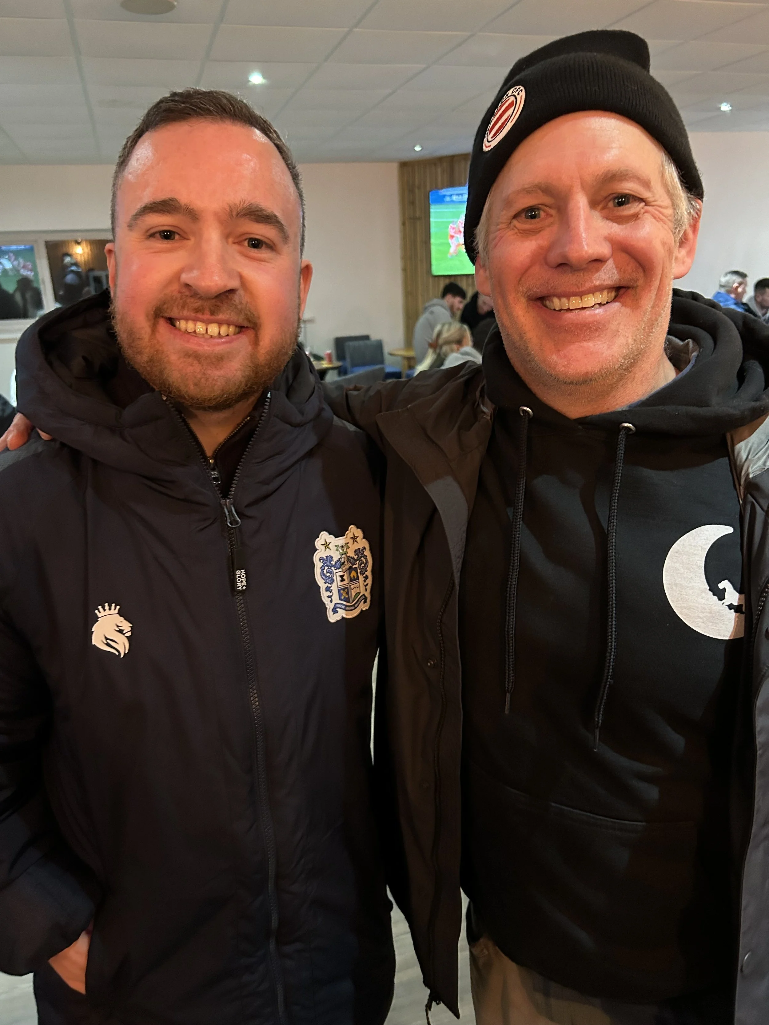Two men smiling and posing for a photo indoors, with a TV screen in the background showing a sports game.
