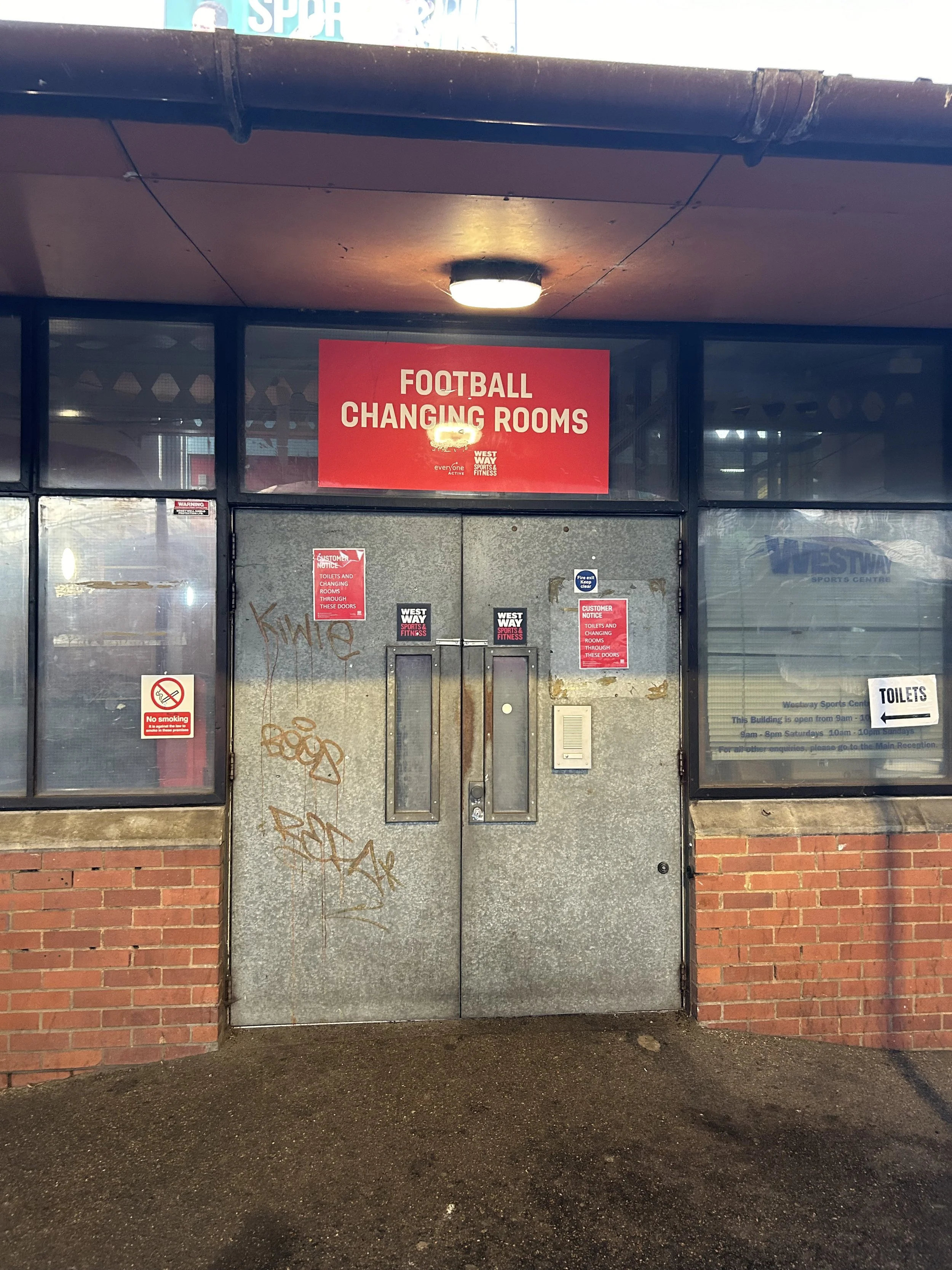 The entrance to football changing rooms with a large red sign above the door that reads 'FOOTBALL CHANGING ROOMS.' The door is metal with graffiti, and there are several smaller notices and signs around it, including a no smoking sign and notices abo